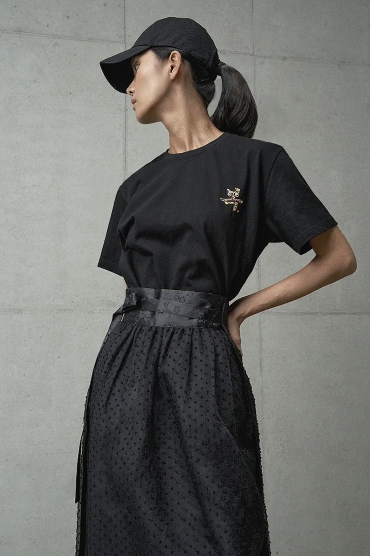 Woman wearing black cap, black t-shirt with floral logo, and textured black skirt against gray wall