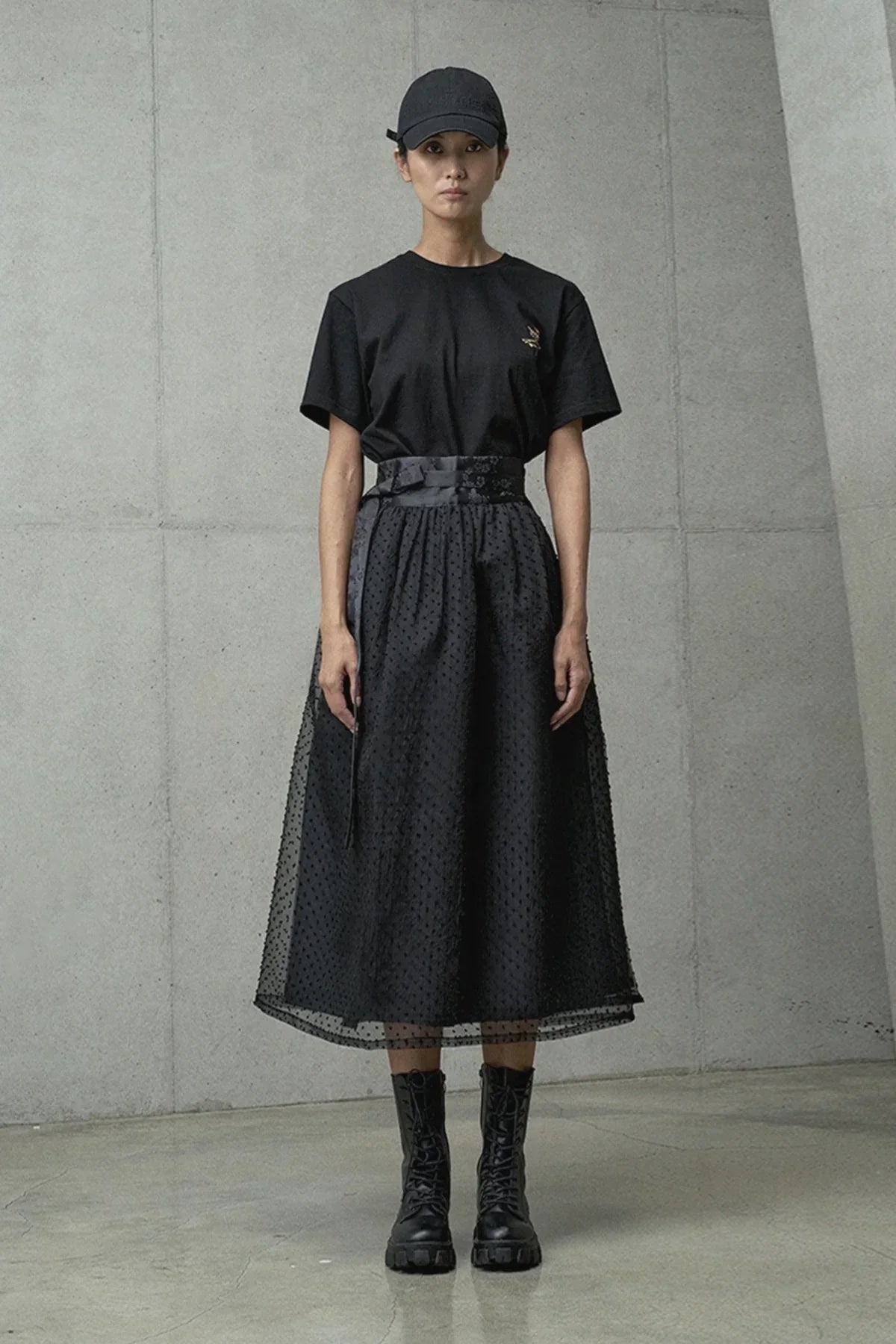 Female model wearing black graphic t-shirt, textured black midi skirt, lace-up boots, and black cap indoors