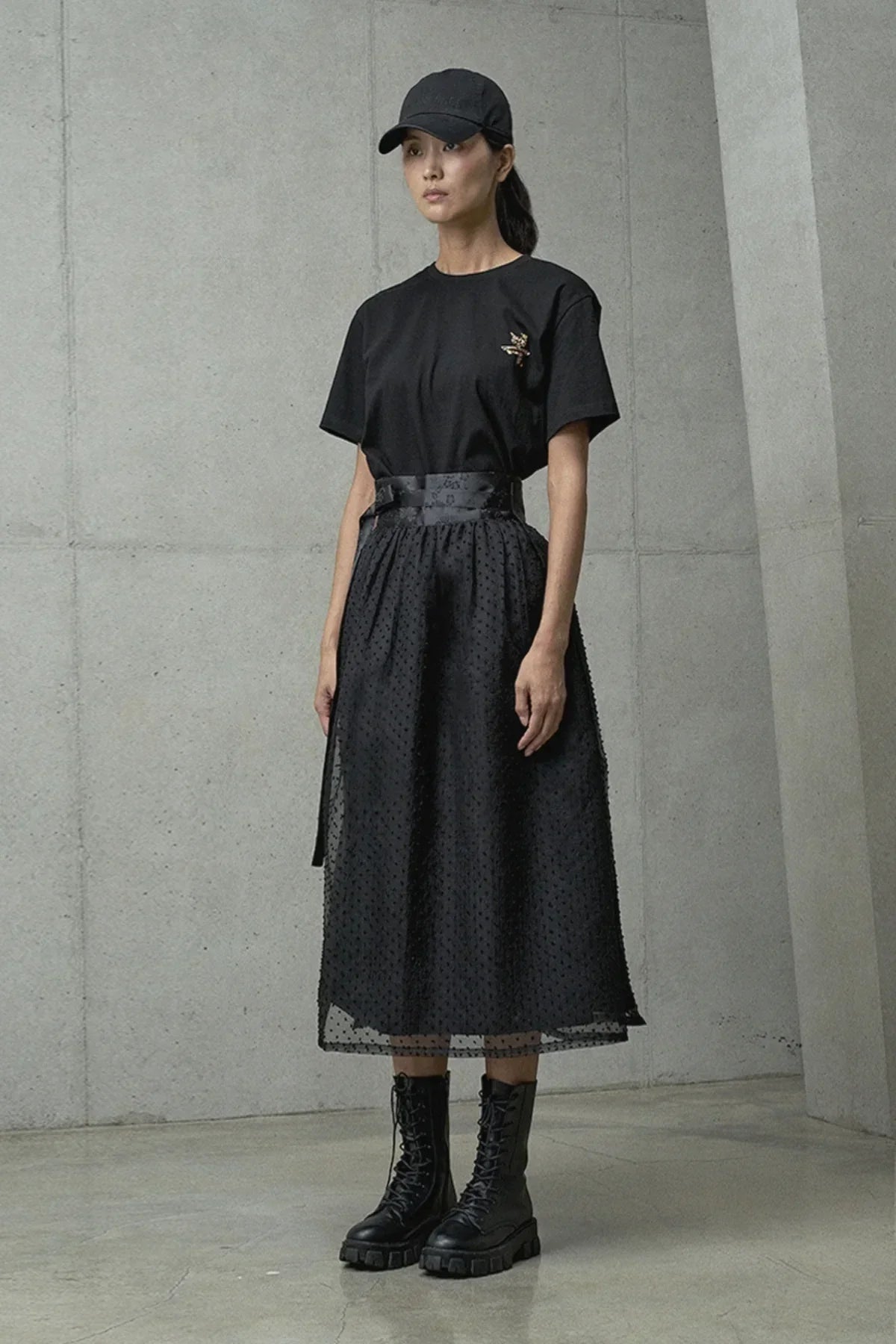 Woman dressed in black cap, embroidered black t-shirt, textured midi skirt, and black lace-up combat boots