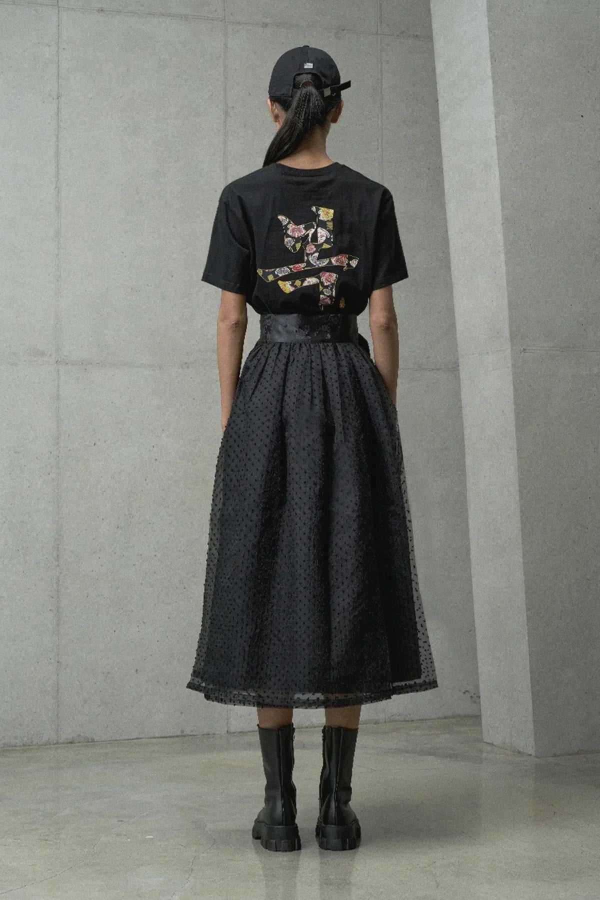 Woman wearing black floral print graphic t-shirt, black textured midi skirt, black cap, and black combat boots standing against concrete wall