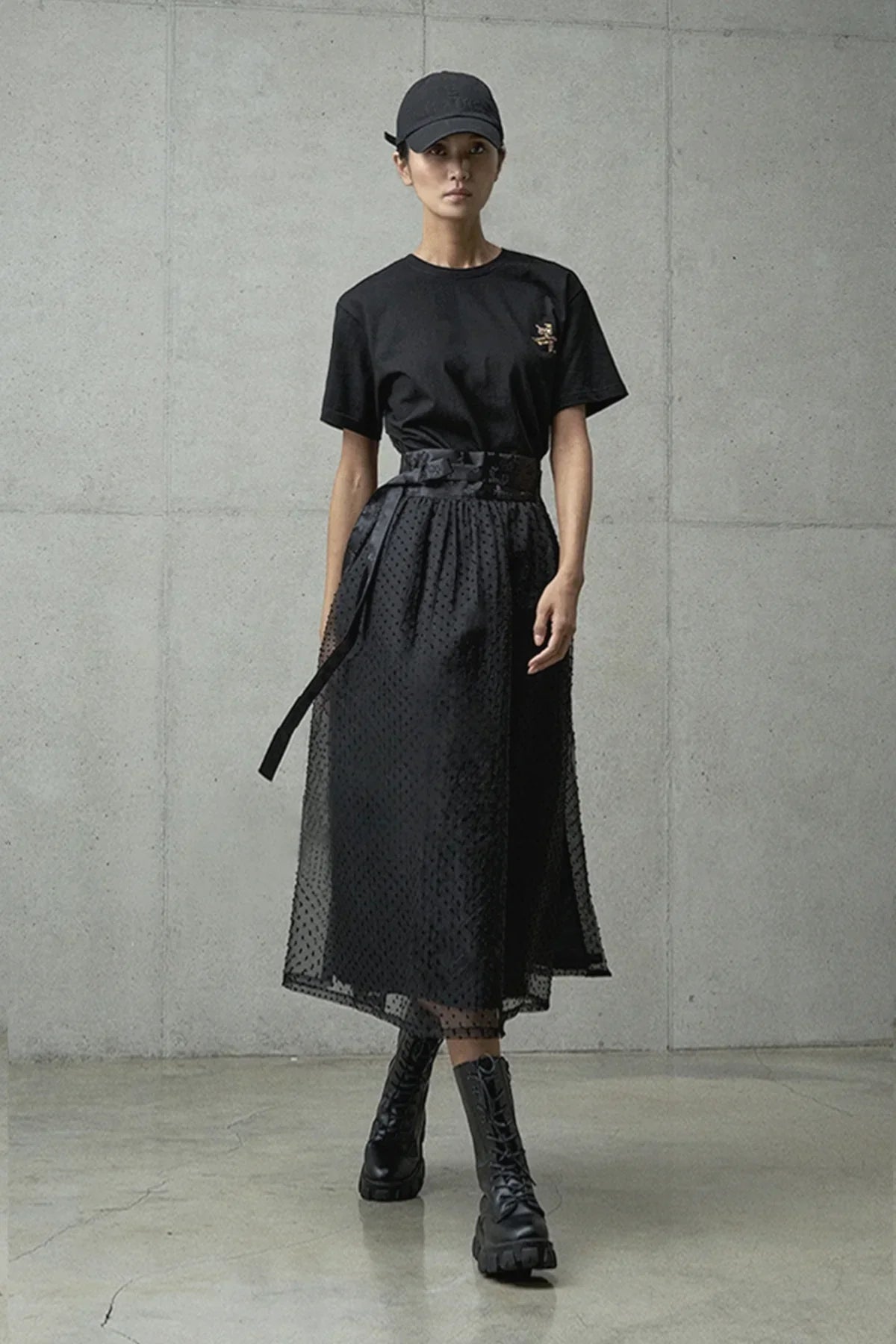 Woman wearing black graphic t-shirt, sheer polka dot midi skirt, black cap, and lace-up combat boots in gray studio