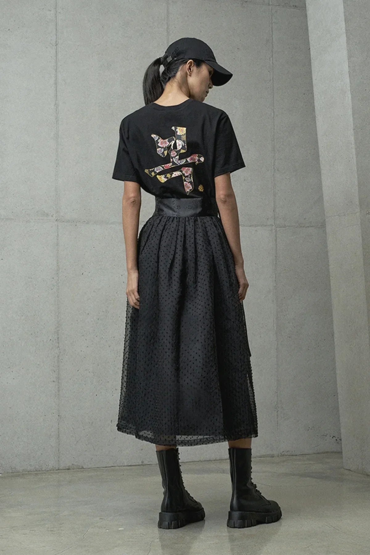 Woman wearing black floral graphic t-shirt, sheer black midi skirt, black boots, and black cap in studio