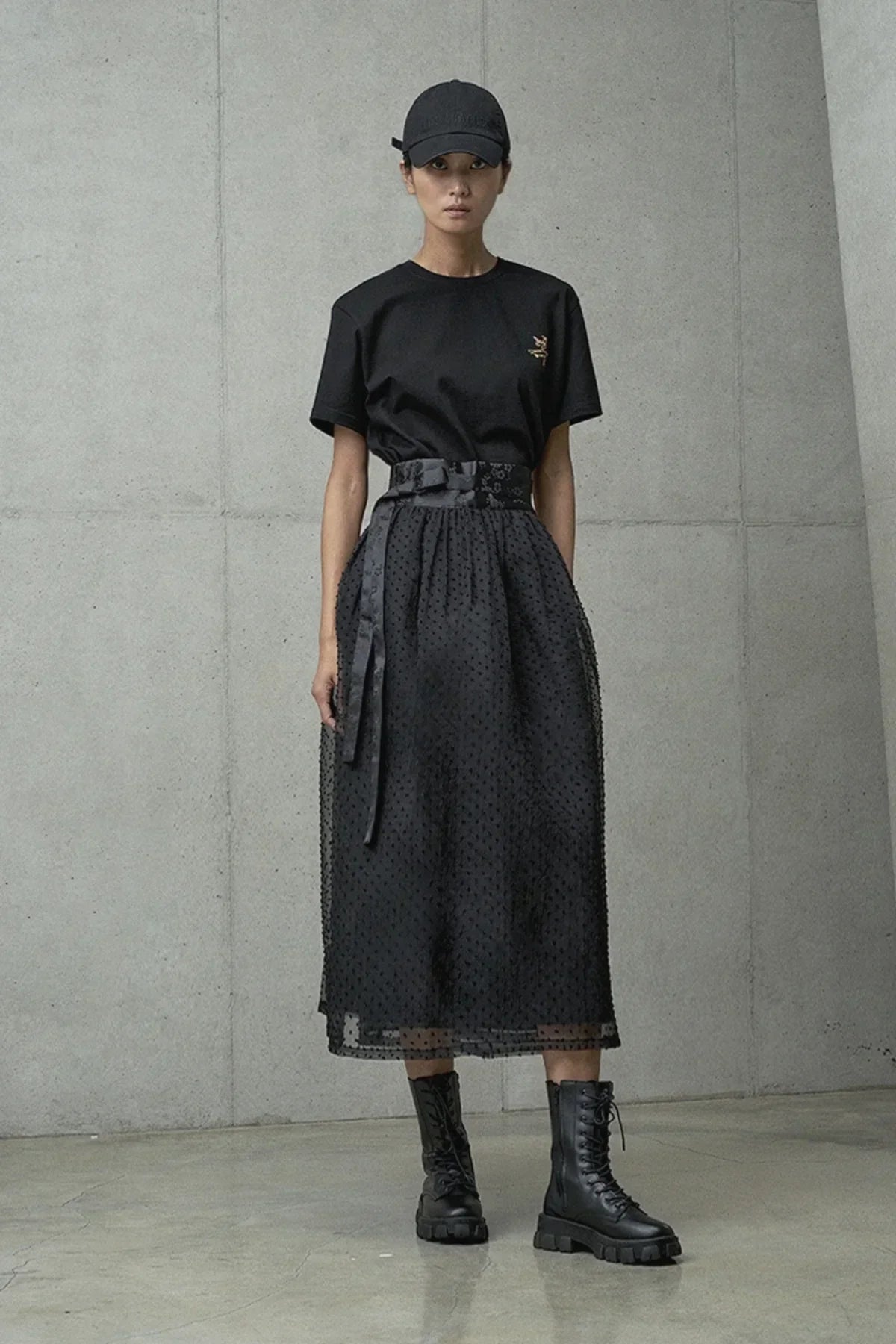 Female model in black cap, graphic t-shirt, sheer polka dot midi skirt, and black lace-up boots on concrete background