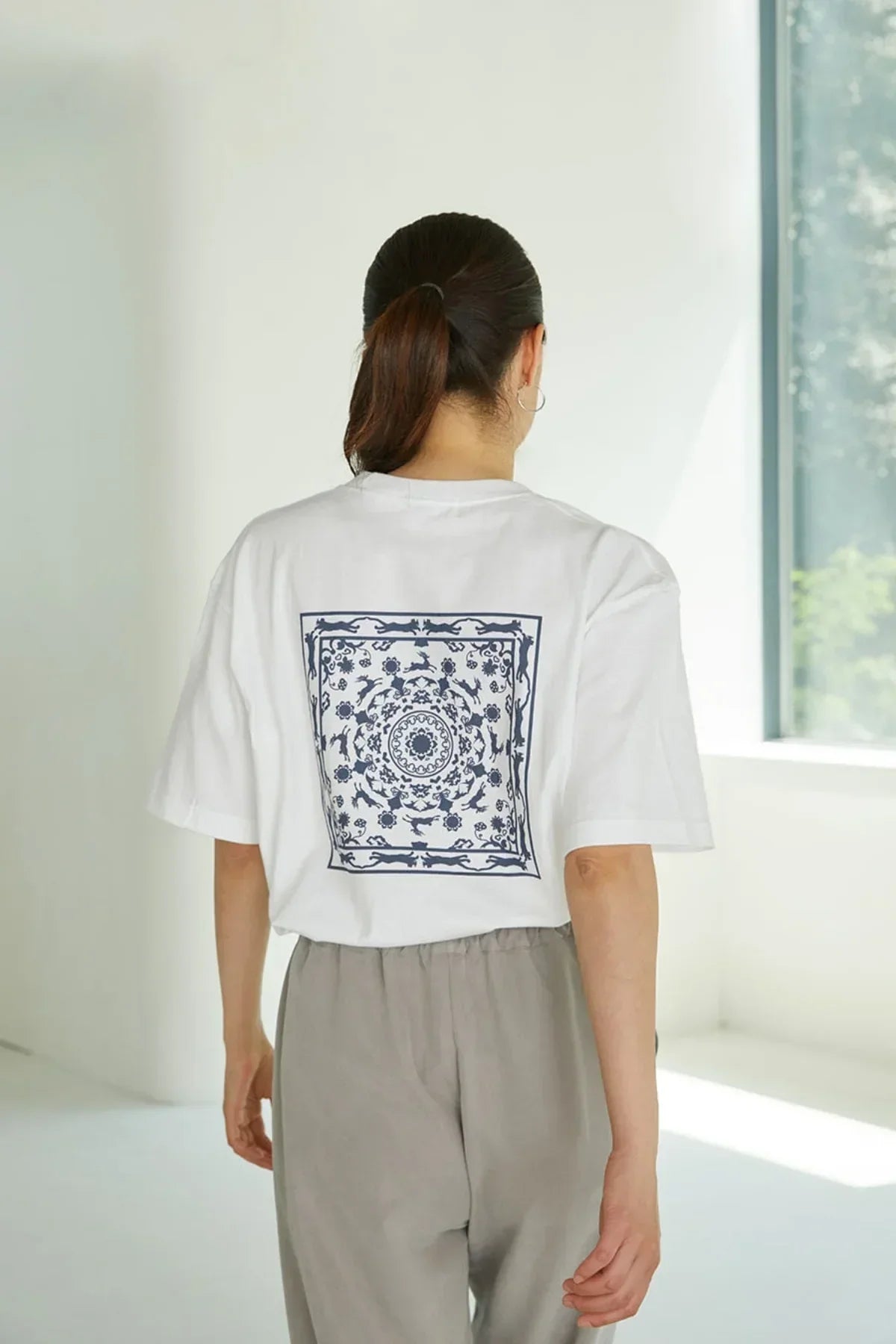 Woman wearing white T-shirt with navy blue intricate circular floral and animal pattern on back, gray pants