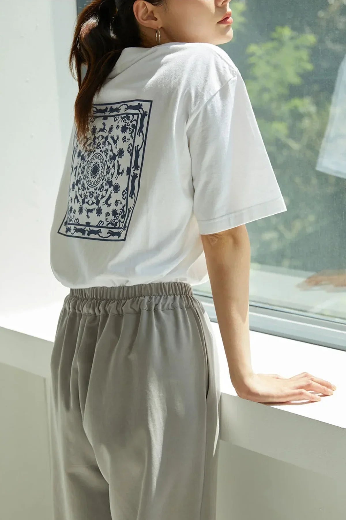 Woman wearing white t-shirt with intricate navy blue Bongsan mask dance print on back, standing by window