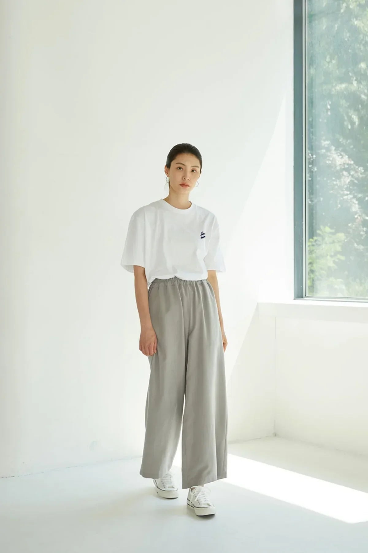 Woman wearing white graphic t-shirt and gray wide-leg pants standing in a bright minimalist room