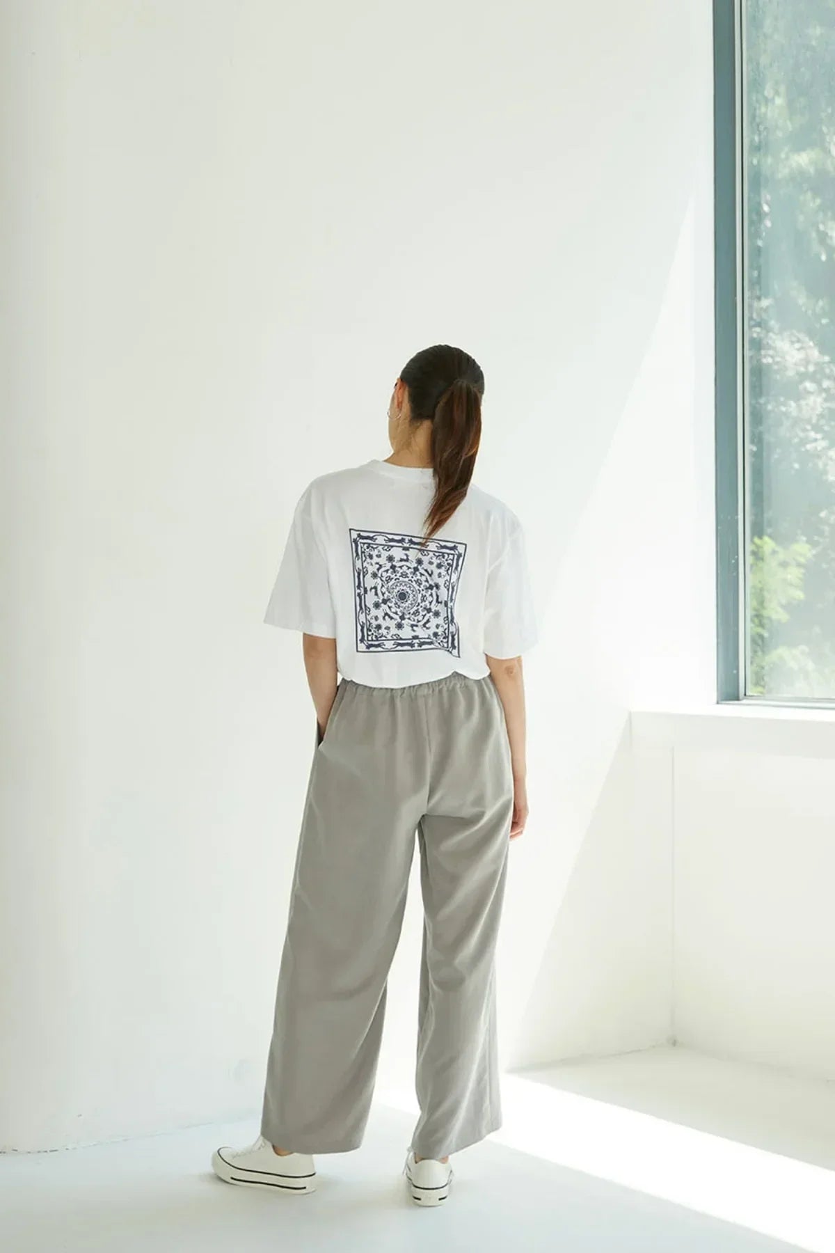 Woman standing in bright room wearing white T-shirt with blue intricate back print and loose gray pants