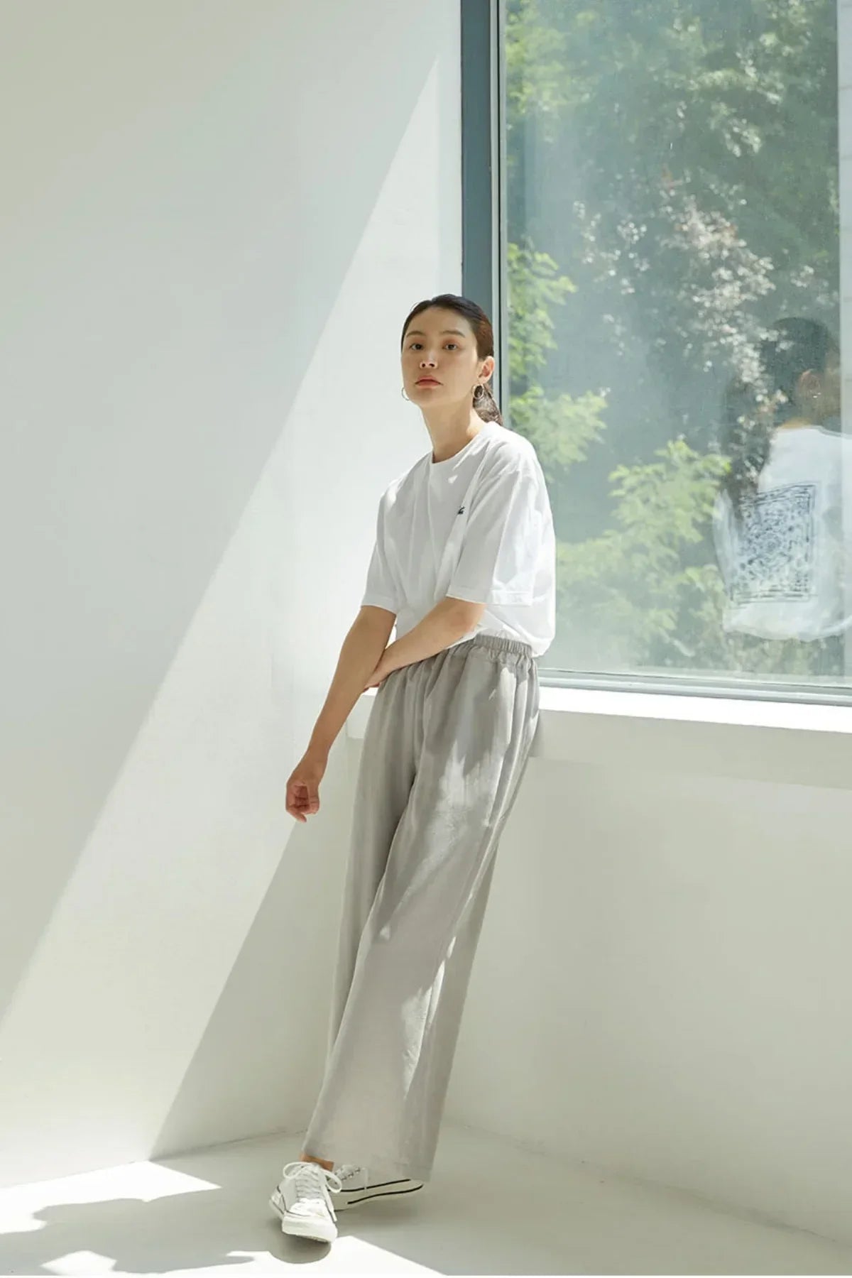 Woman in white t-shirt and light gray wide-leg pants standing near window in bright sunlit room