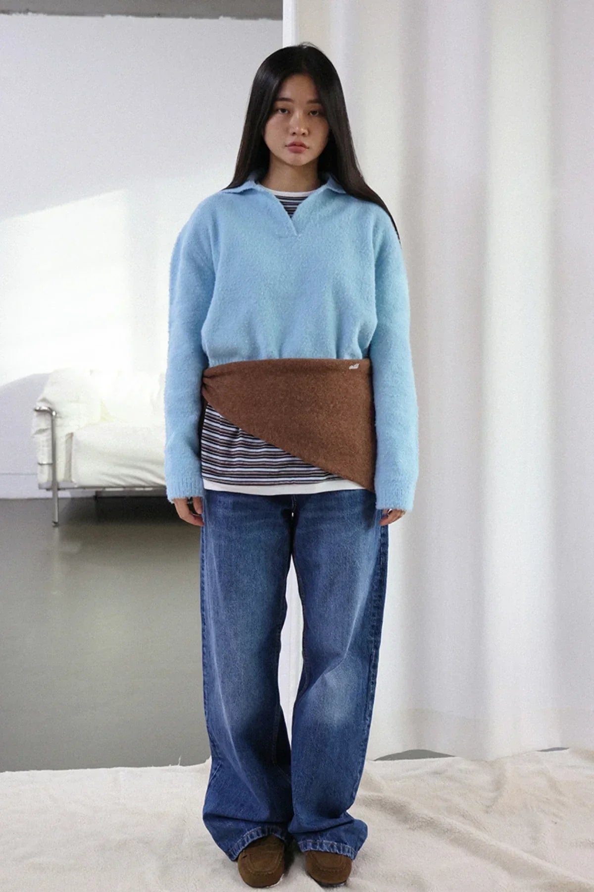 Woman wearing sky blue fuzzy knit sweater, striped shirt, wide jeans, and brown wrap belt in minimalist room
