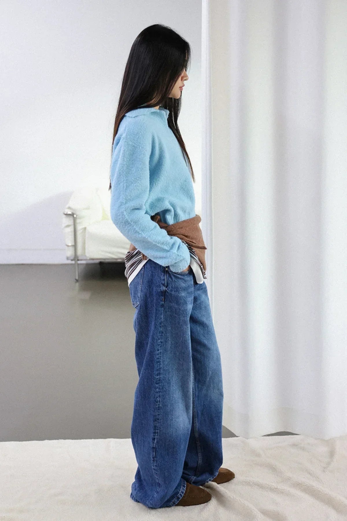 Side view of woman wearing sky blue knit sweater and loose blue jeans in minimalist indoor setting