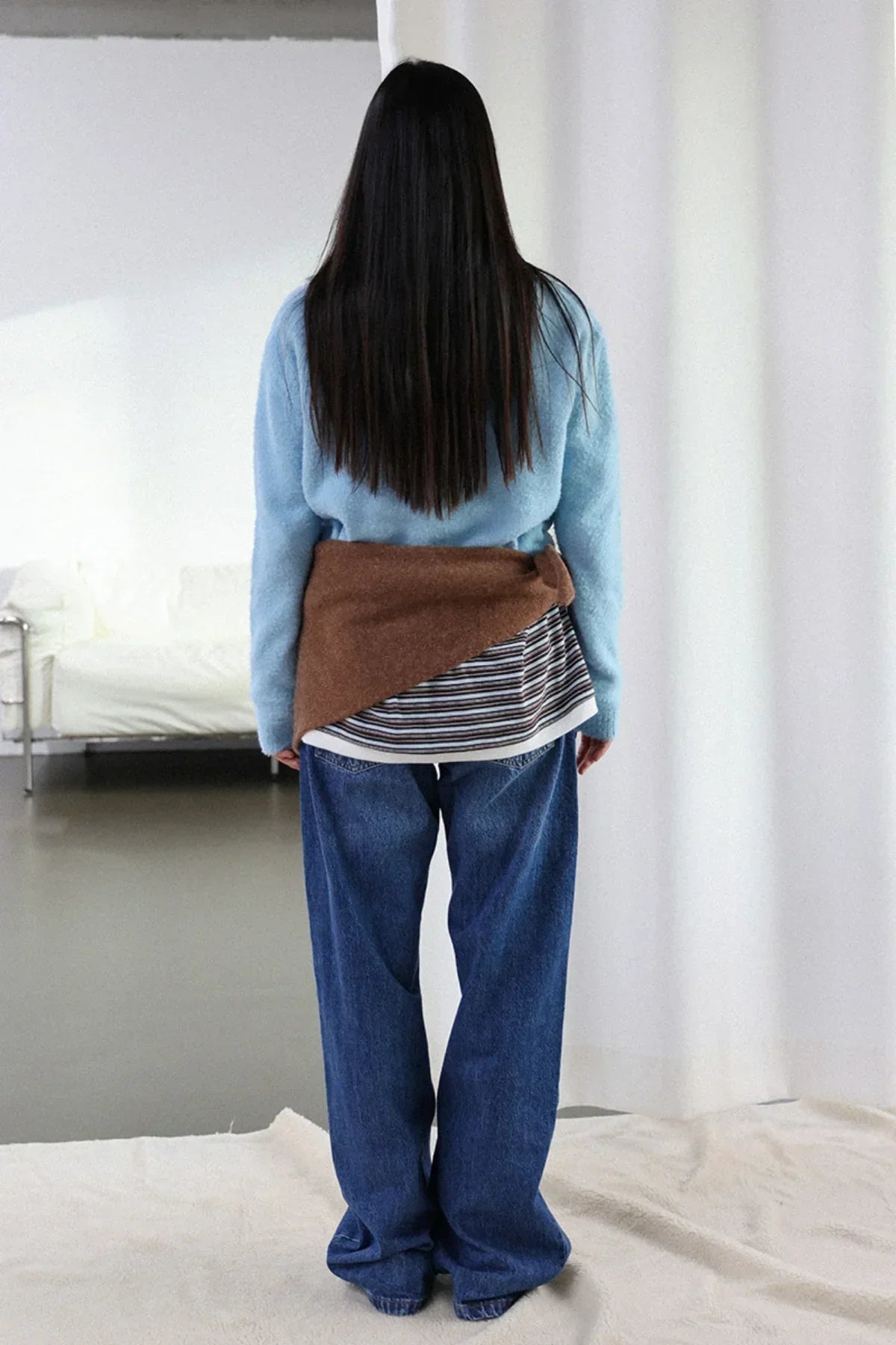 Woman with long dark hair wearing sky blue knit sweater, striped shirt, brown waist wrap, and loose blue jeans indoors