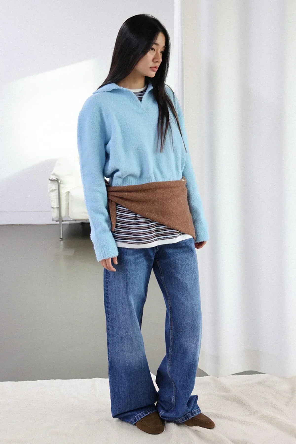 Young woman wearing sky blue knit sweater, striped shirt, wide leg jeans, and brown shoes in minimalist studio