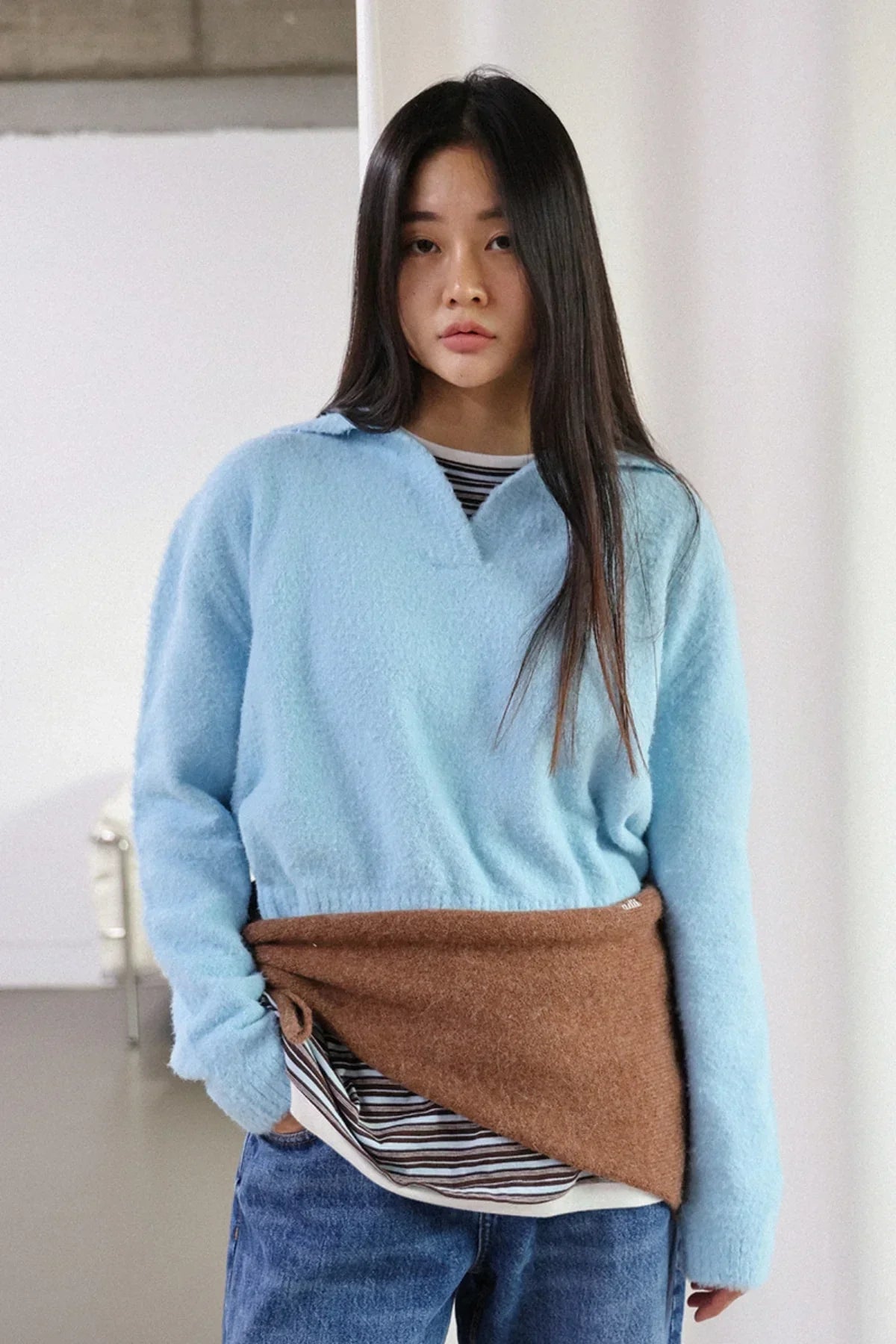 Young woman wearing sky blue fuzzy knit sweater, brown wrap skirt, and striped shirt in minimal studio