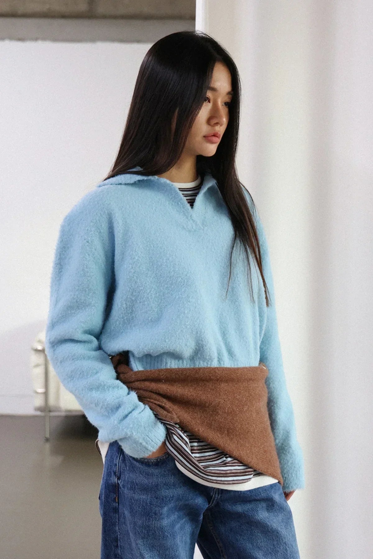 Young woman wearing sky blue fuzzy knit sweater, brown waist wrap, striped shirt, and blue jeans in studio