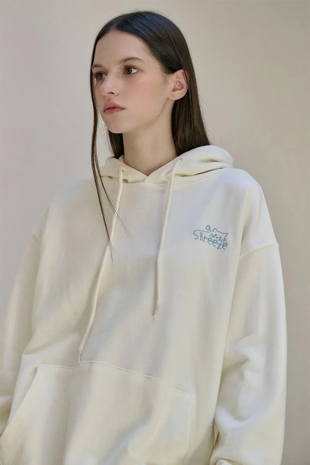 Young woman wearing cream hoodie with blue embroidered cat and breeze logo detail