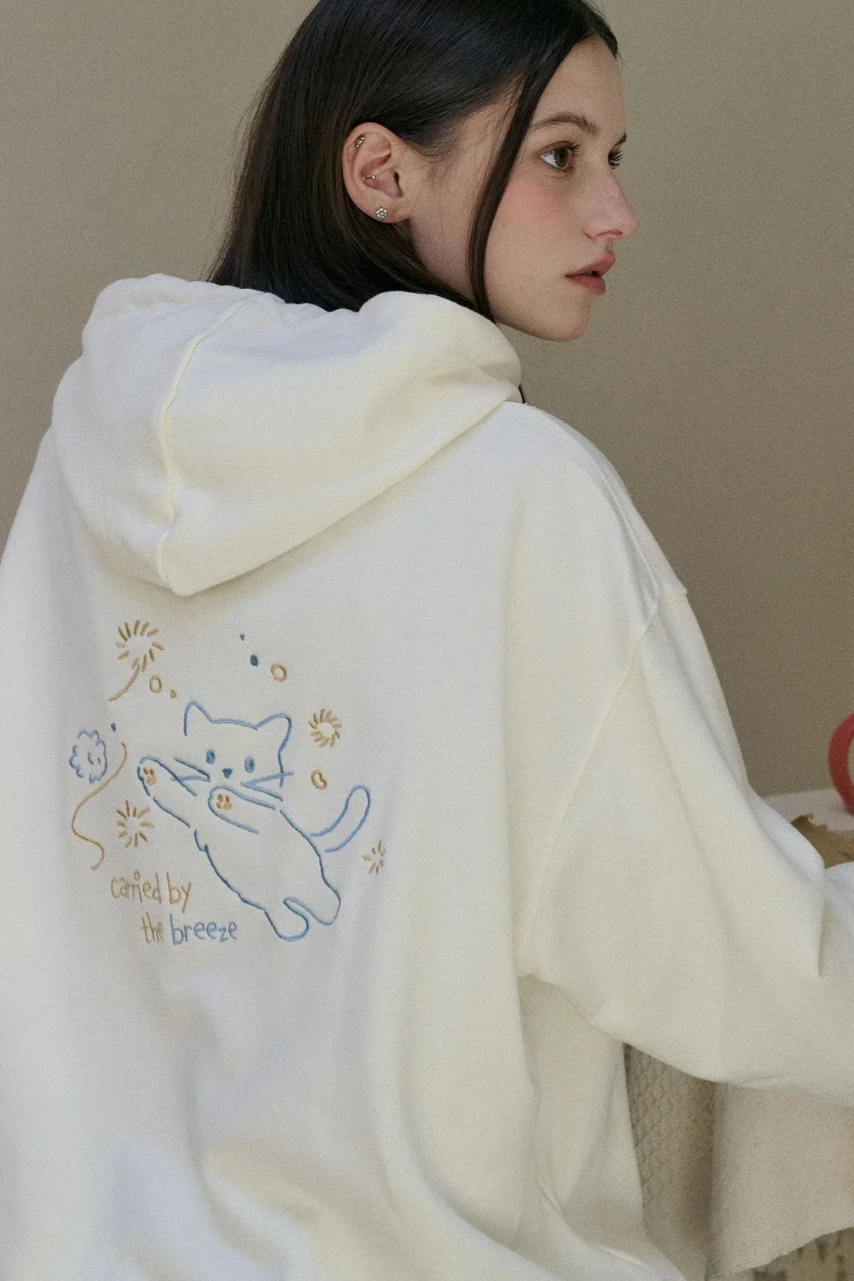 Young woman in cream hoodie with blue cat embroidery and text 'carried by the breeze' on back