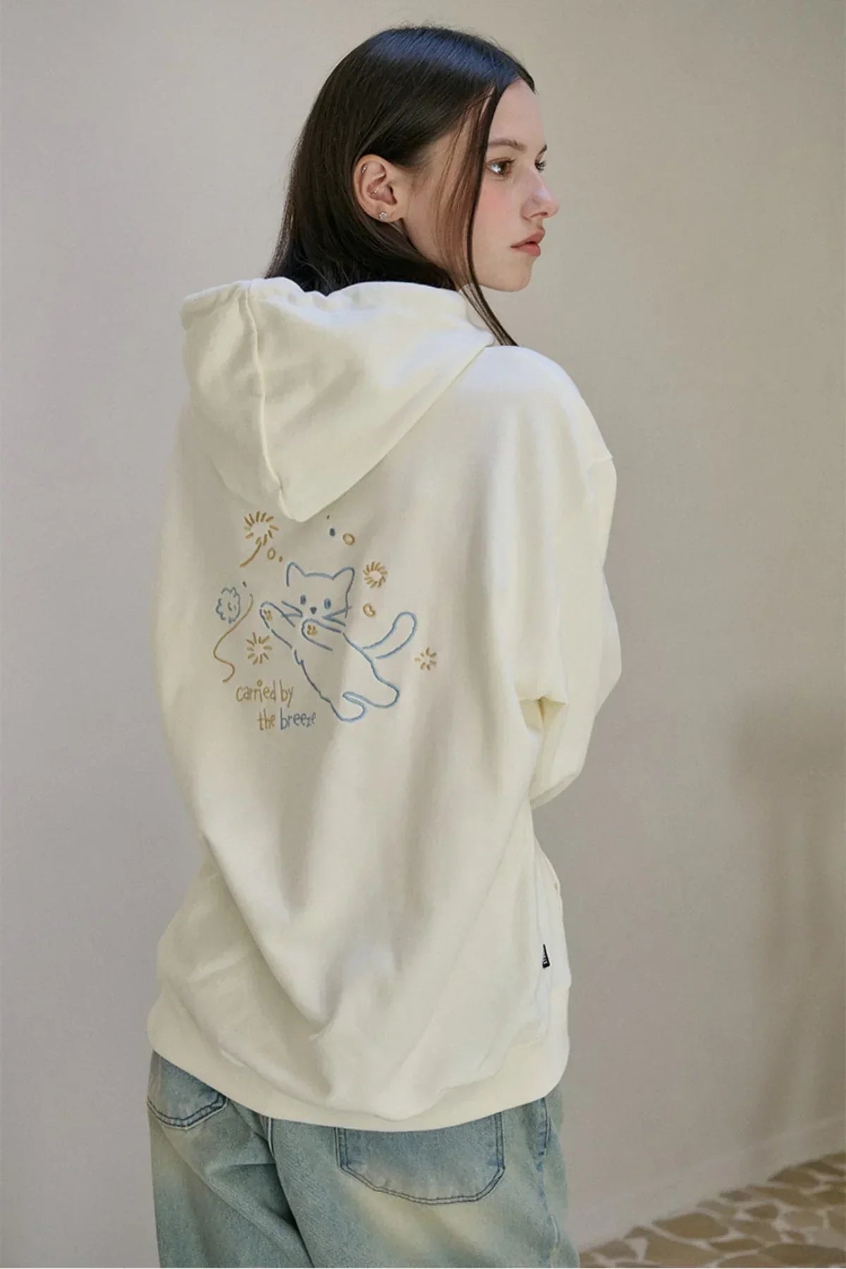 Woman wearing a cream hoodie with an embroidered playful cat and the text carried by the breeze on the back