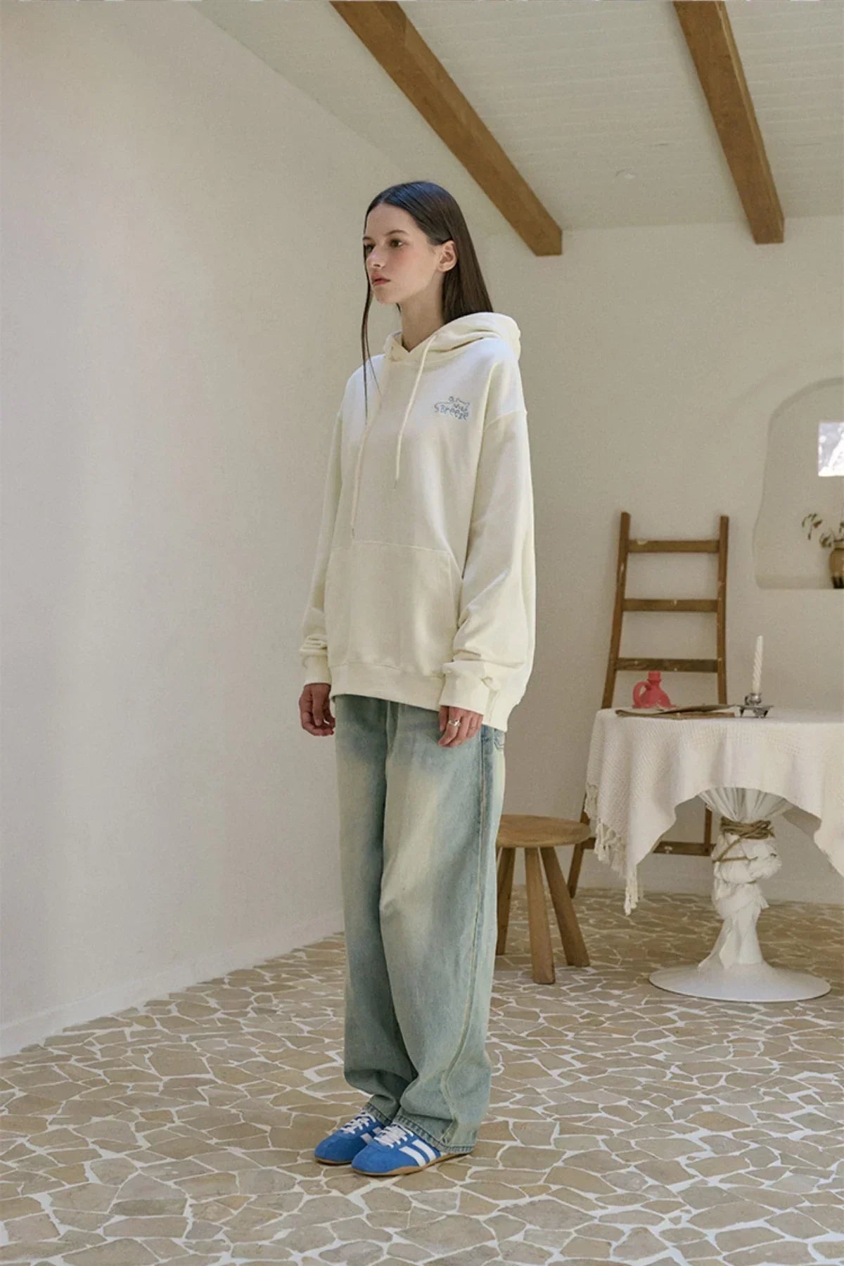 Woman wearing cream BreezeCat embroidered hoodie with light wash jeans and blue sneakers in rustic room