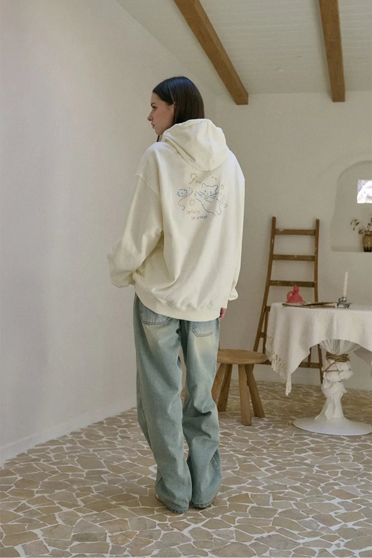Woman wearing cream embroidered hoodie with cat design and loose blue jeans in cozy room