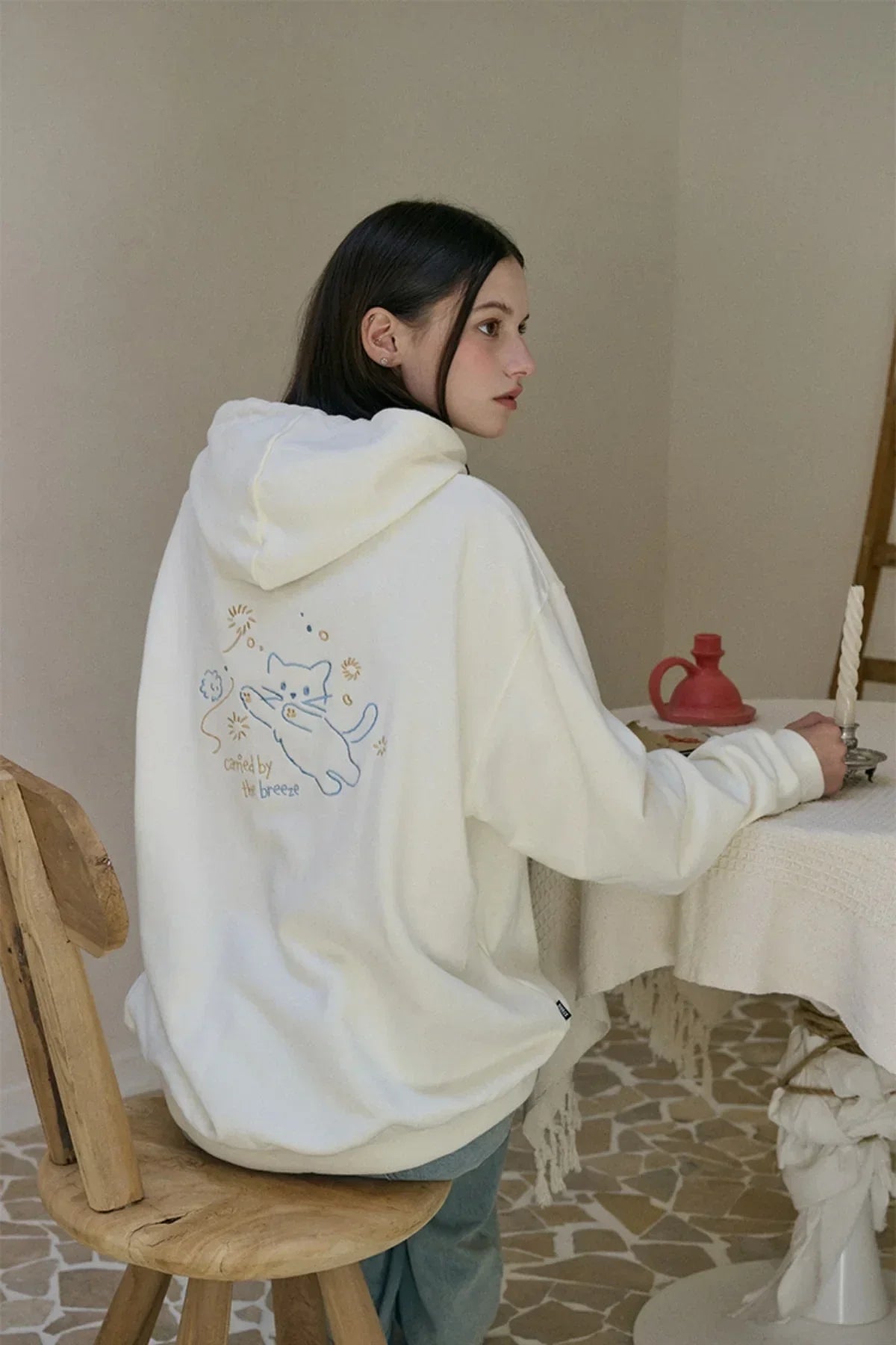 Woman wearing cream hoodie with embroidered playful cat and text sitting on wooden stool indoors