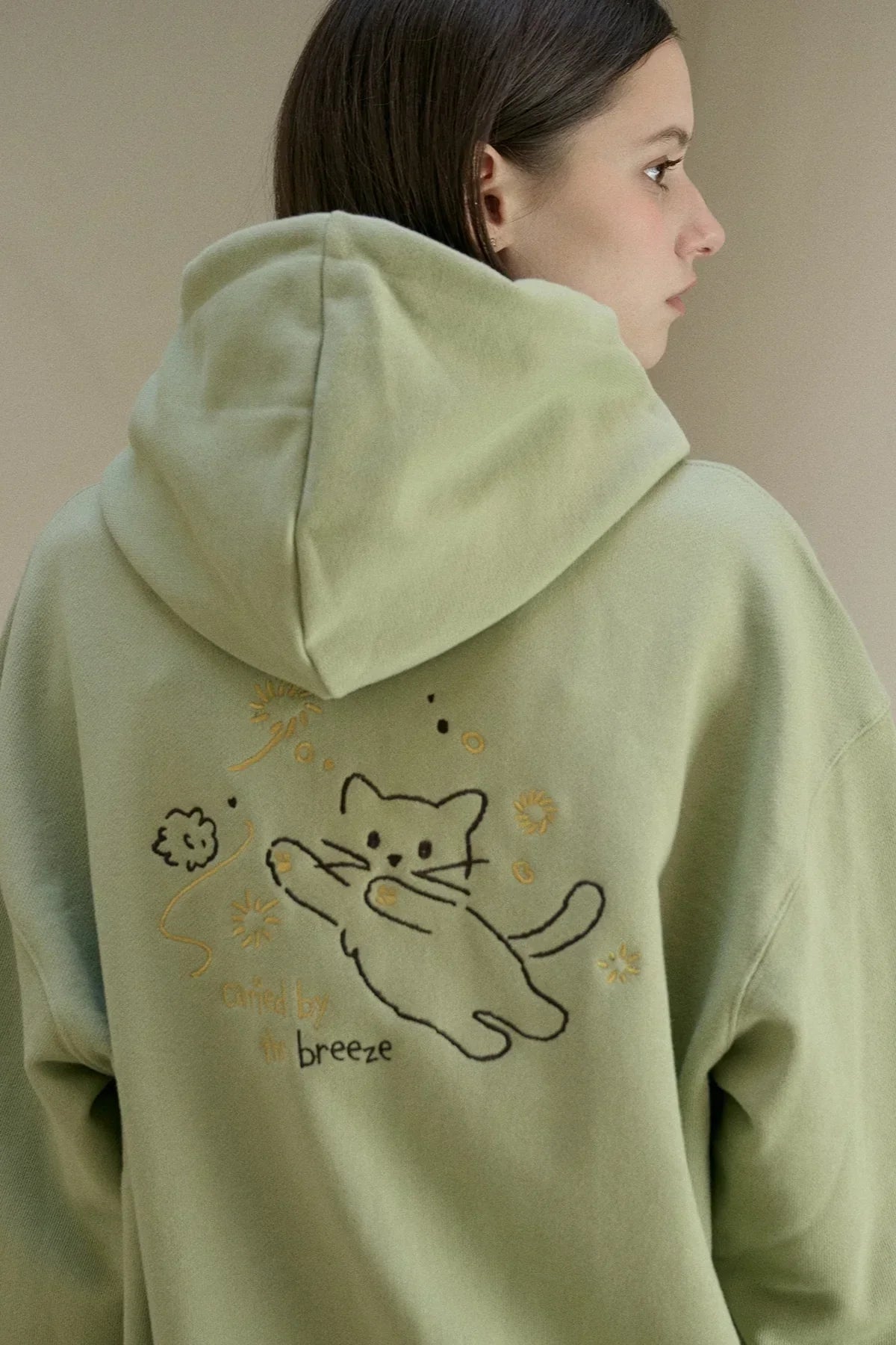 Side view of woman wearing light green hoodie with embroidered playful cat and decorative text on back