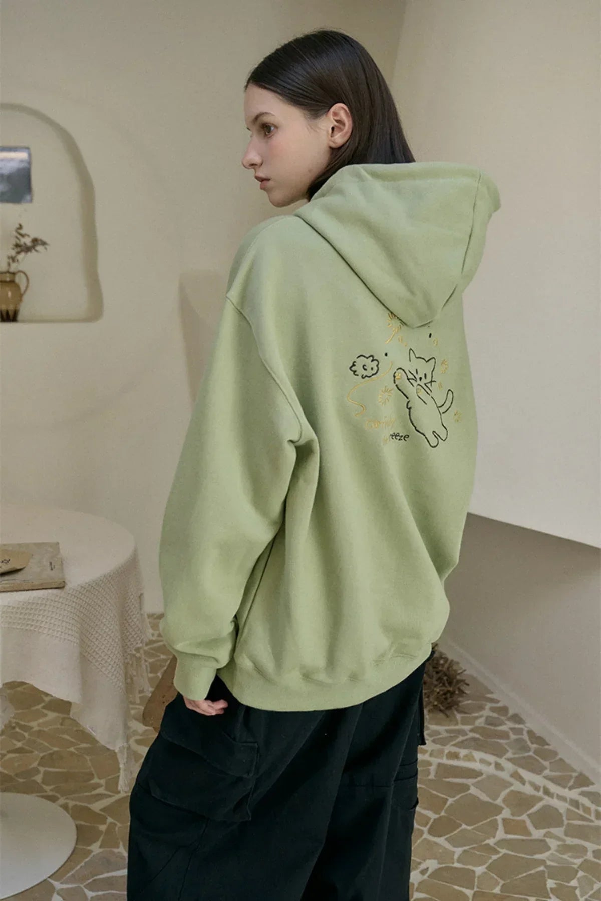 Woman wearing khaki hoodie with embroidered cat design on back, standing in minimalist room
