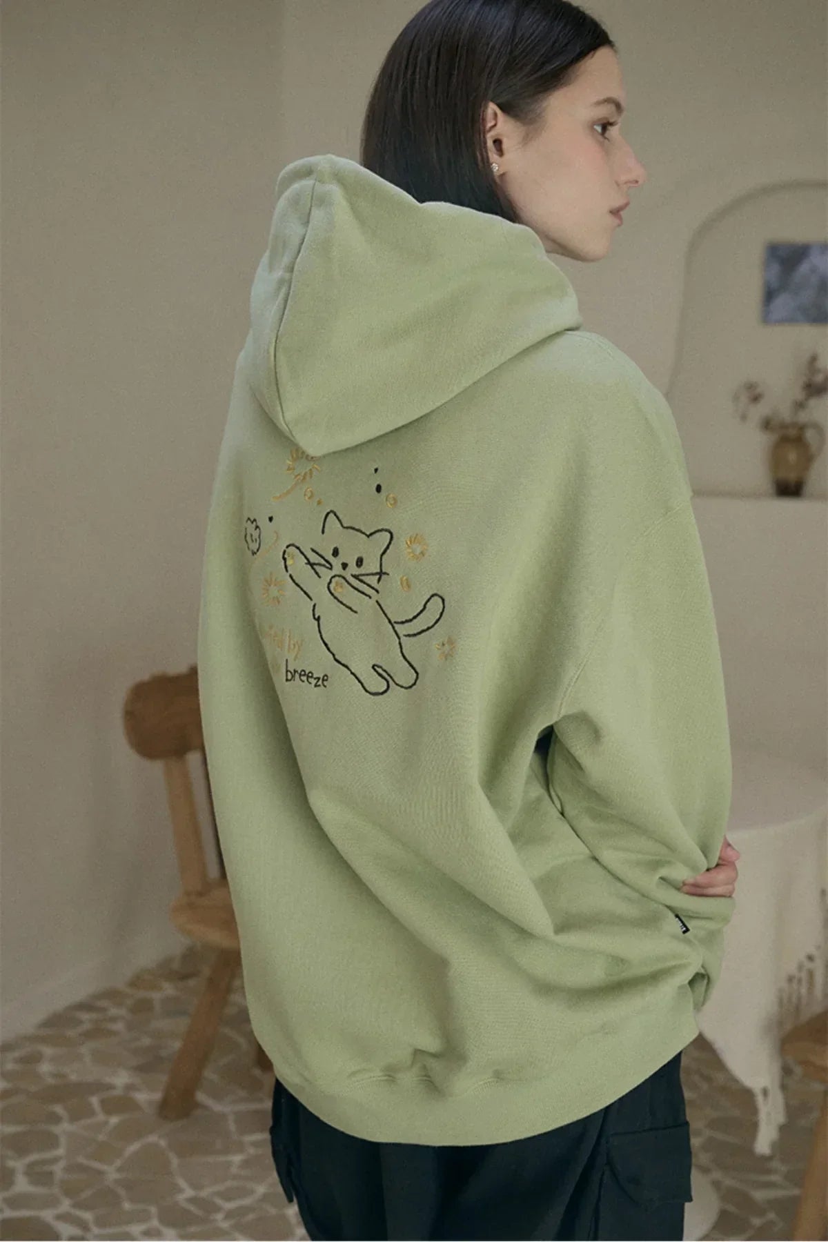 Person wearing khaki oversized hoodie with embroidered playful cat design on back indoors