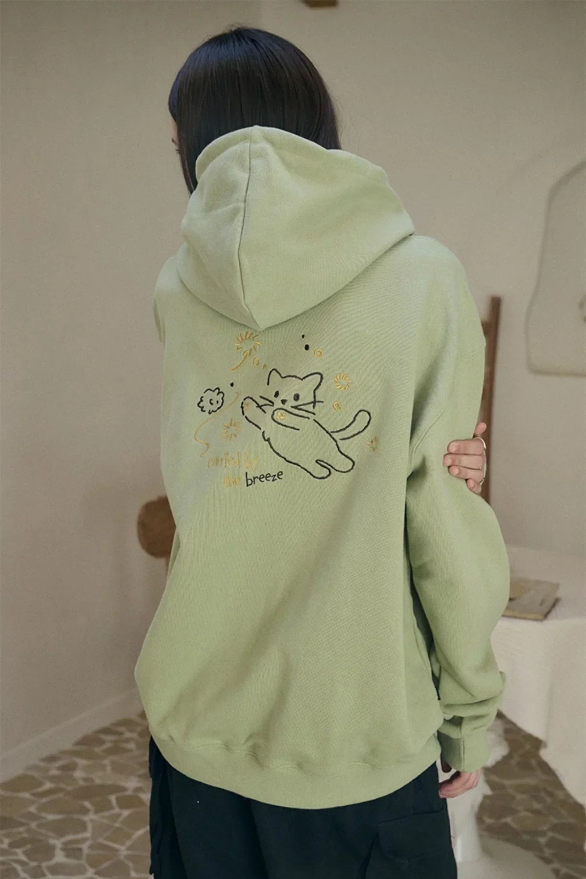 Back view of person wearing light green hoodie with embroidered cat and text in indoor setting