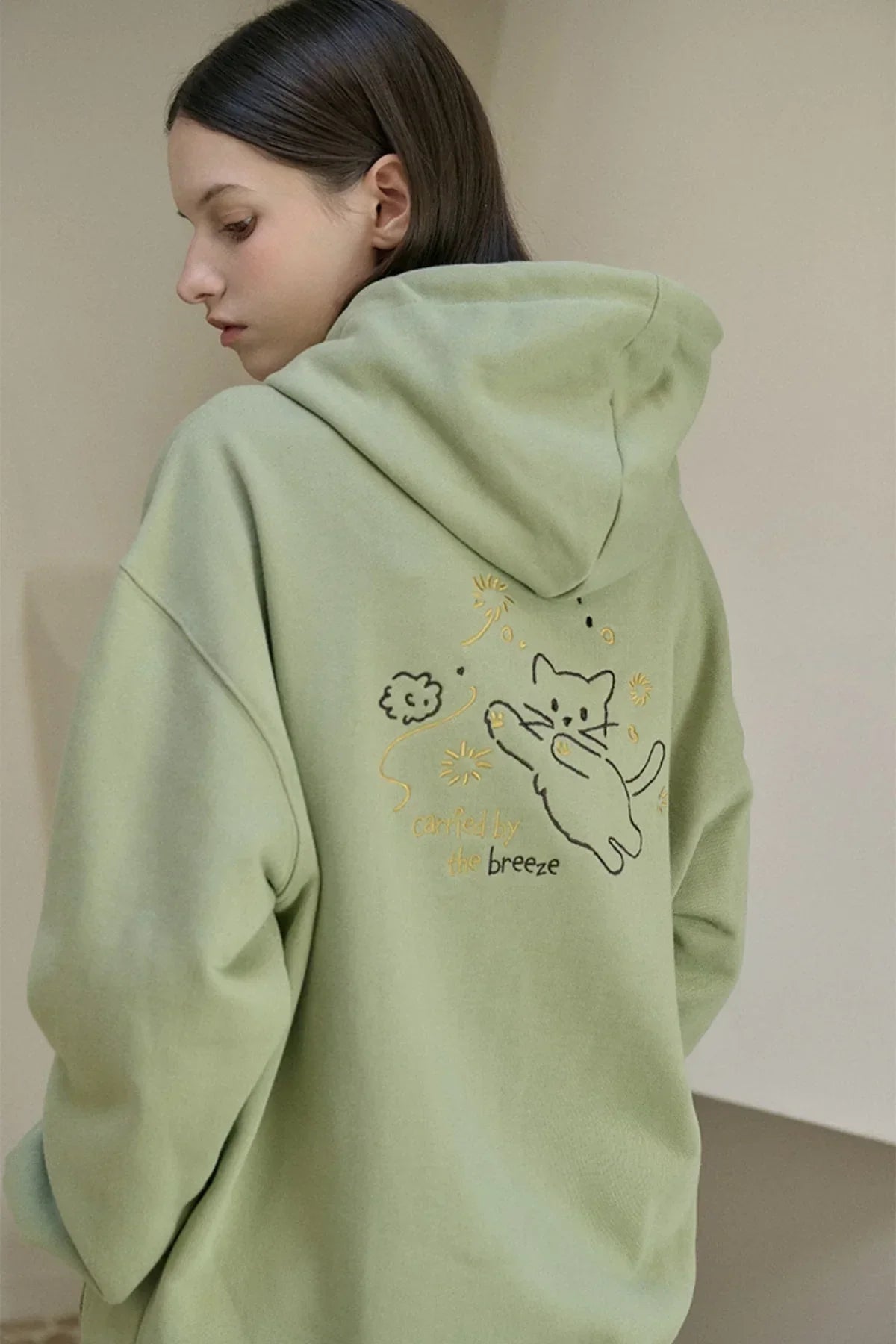 Woman wearing khaki green hoodie with embroidered playful cat and text carried by the breeze