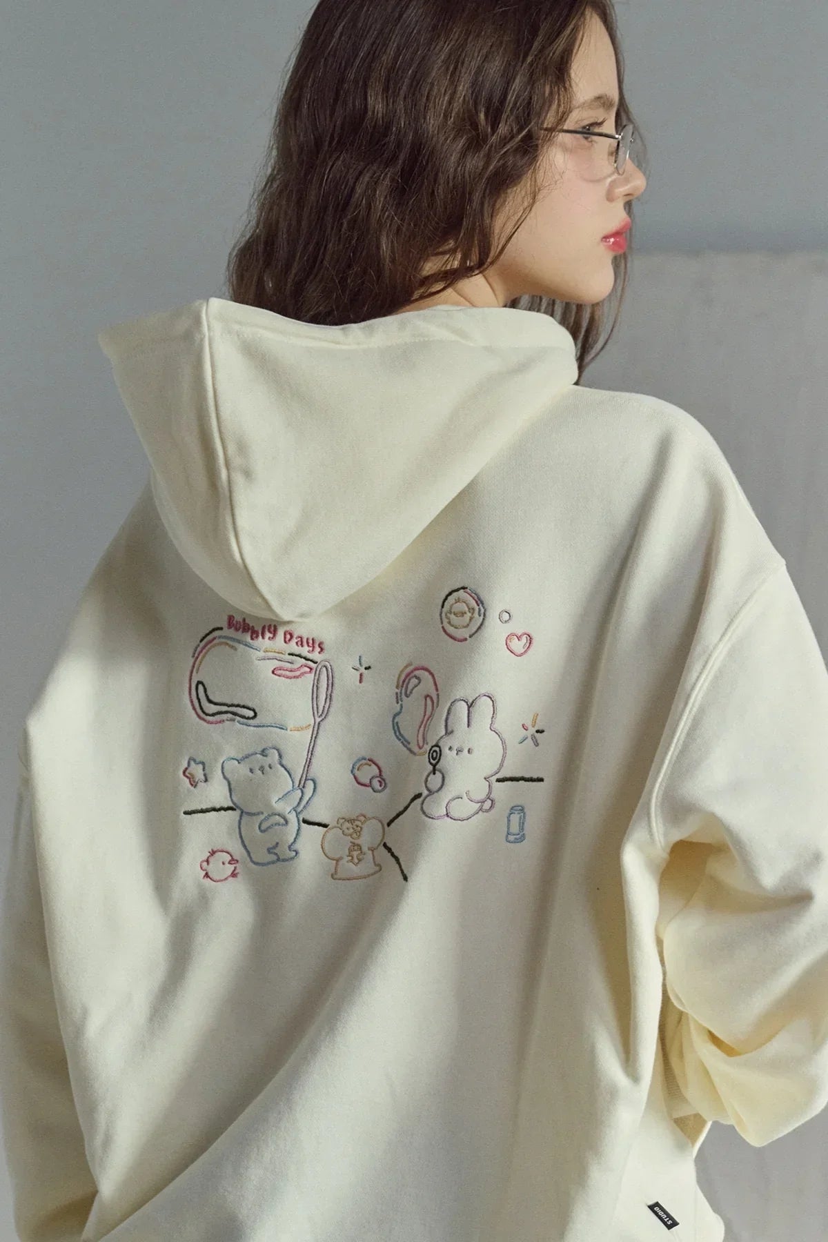 woman wearing cream hoodie with colorful bubbly days embroidered cartoon characters on back