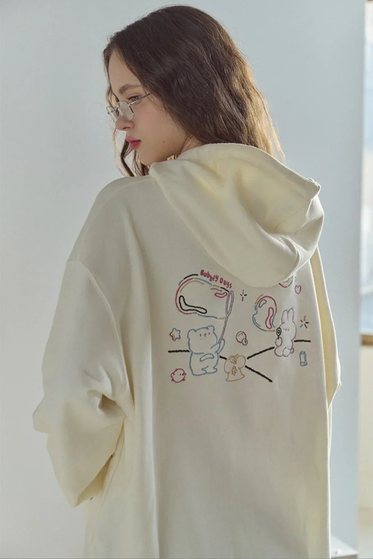 Woman wearing cream hoodie with colorful embroidered cartoon characters and text on back