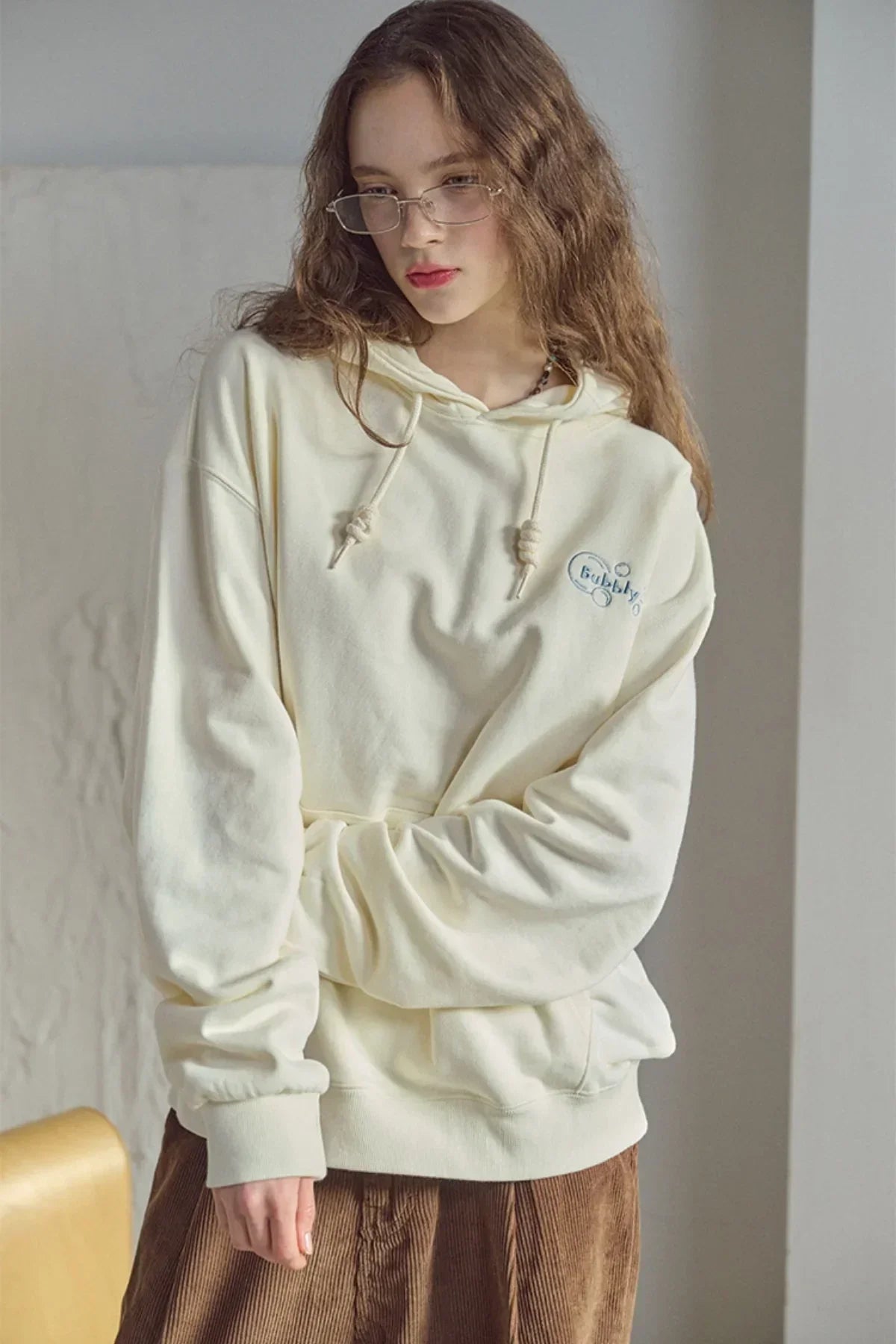Woman wearing cream oversized hoodie with 'Bubbly' embroidery and brown corduroy skirt indoors