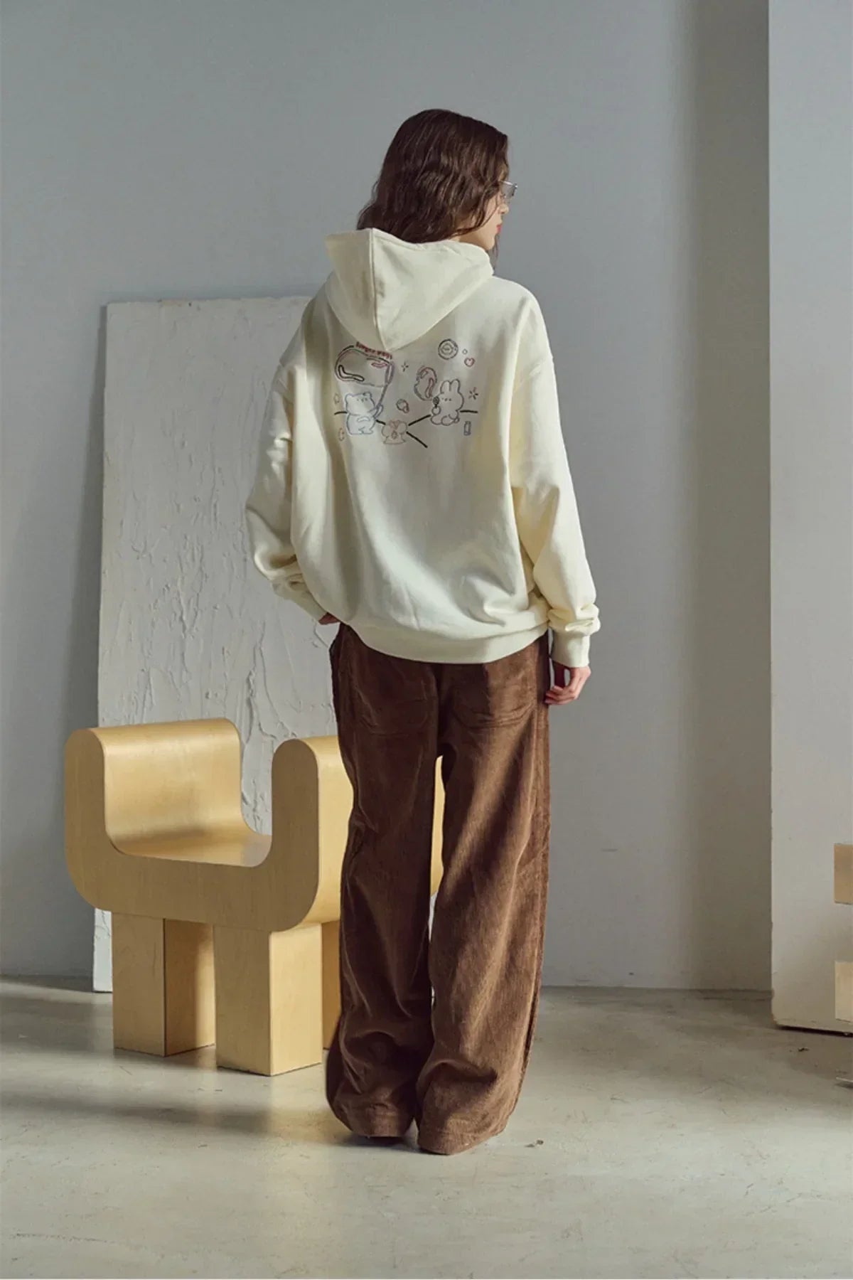 Woman wearing cream hoodie with cute embroidered cartoon design and brown wide-leg pants in minimal room