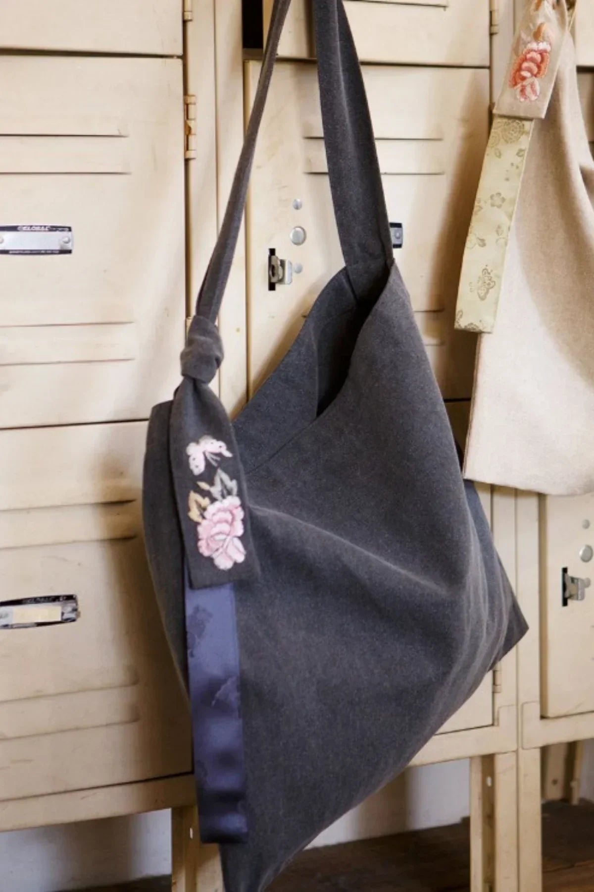 Dark gray fabric tote bag with floral embroidery and wide strap hanging on vintage cream metal lockers