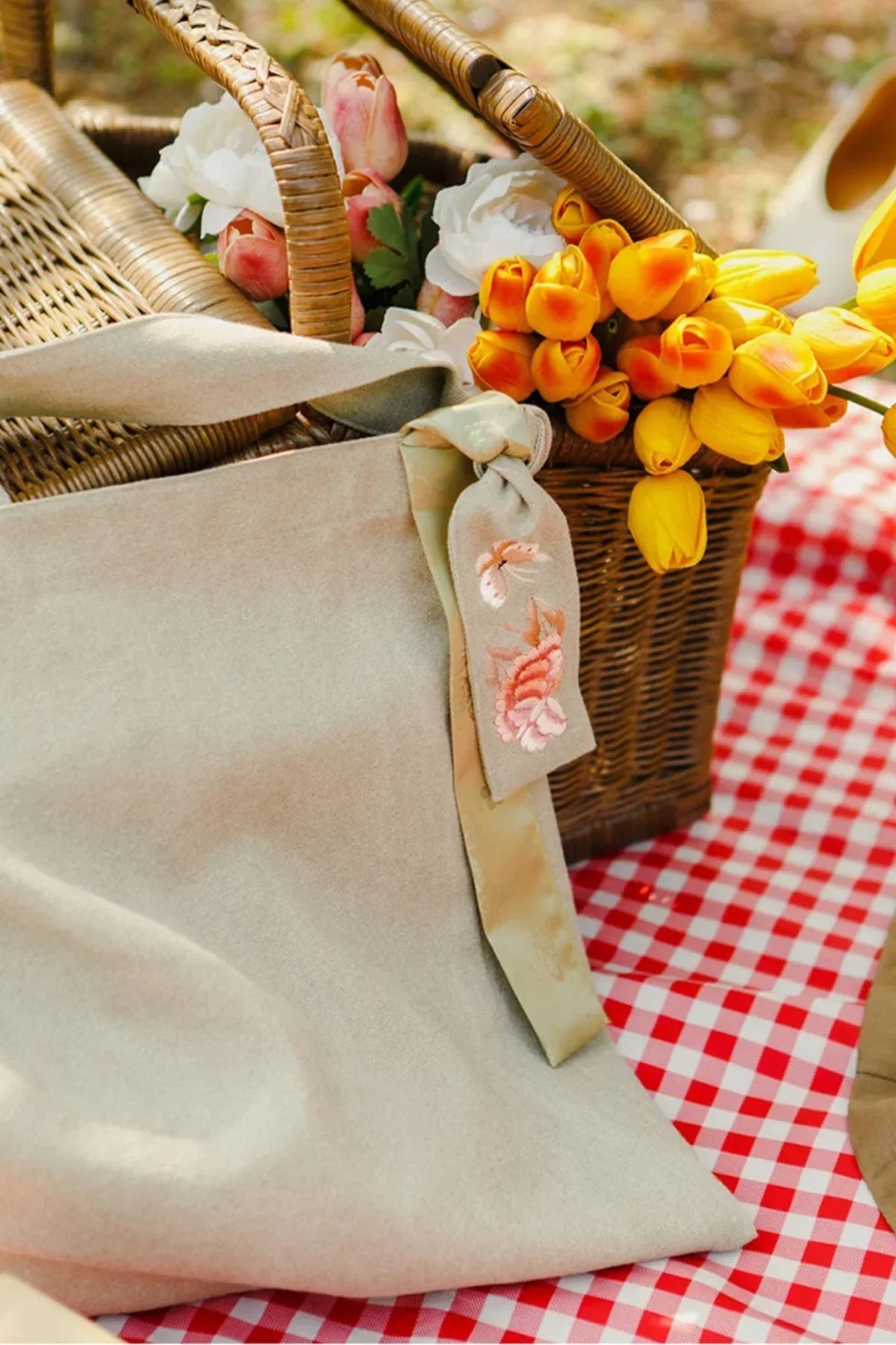 Beige fabric daily bag with embroidered butterfly detail beside wicker basket of mixed tulips on red check picnic blanket