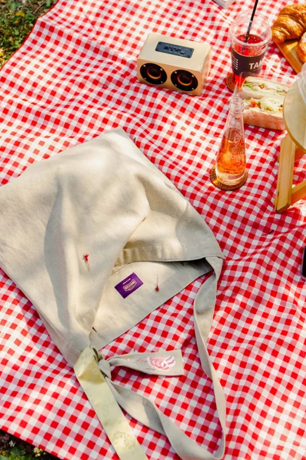 Beige fabric bag on red and white checkered picnic blanket with drinks, sandwich, croissants, and speaker