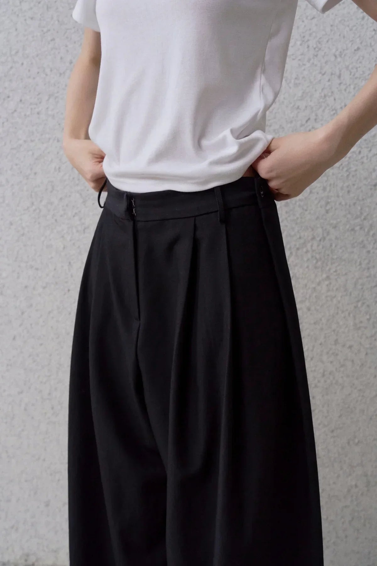 Close-up of person wearing wide fit black pants and white top against a gray textured wall