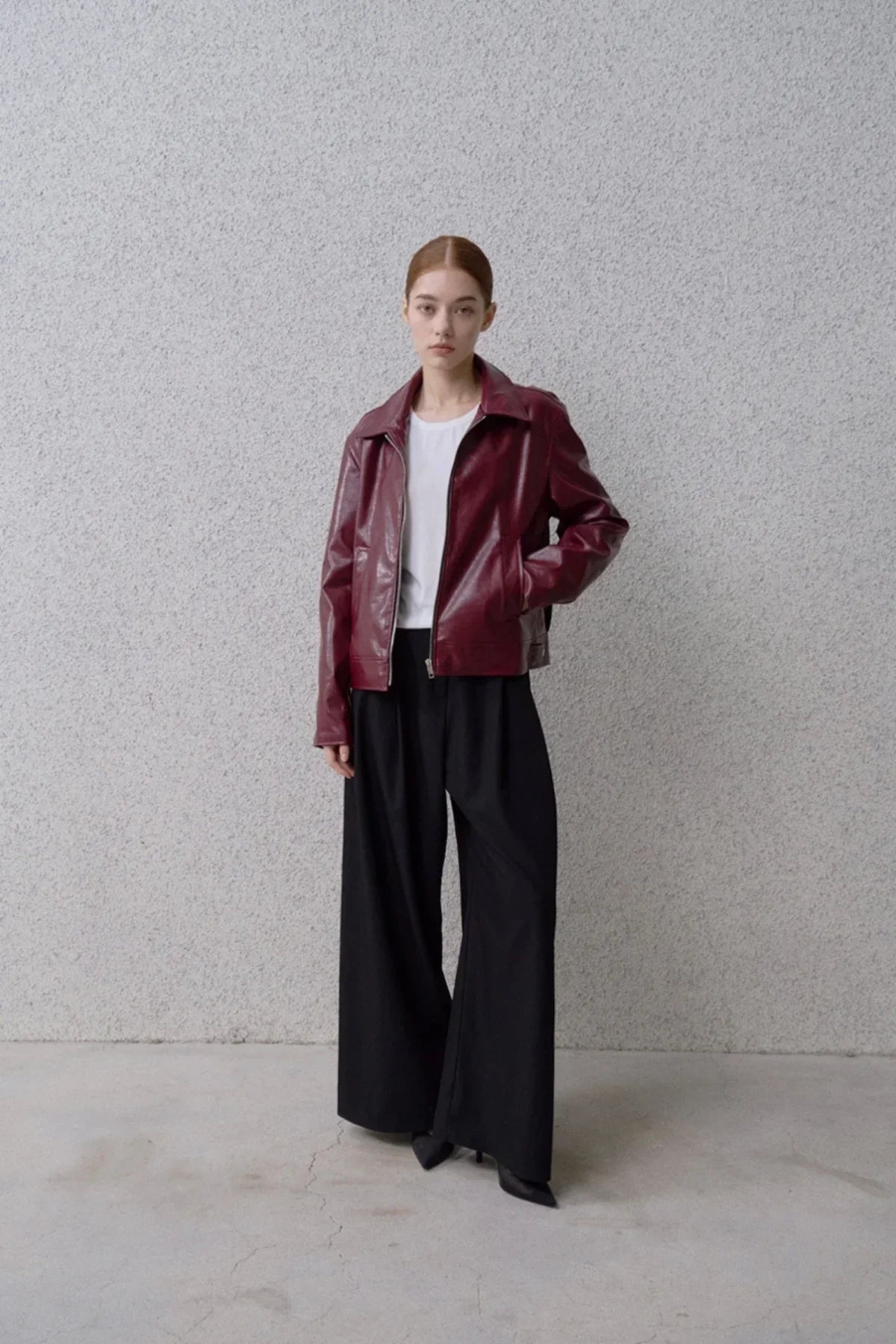 Model wearing black wide-leg pants with a white shirt and burgundy leather jacket standing against grey textured wall