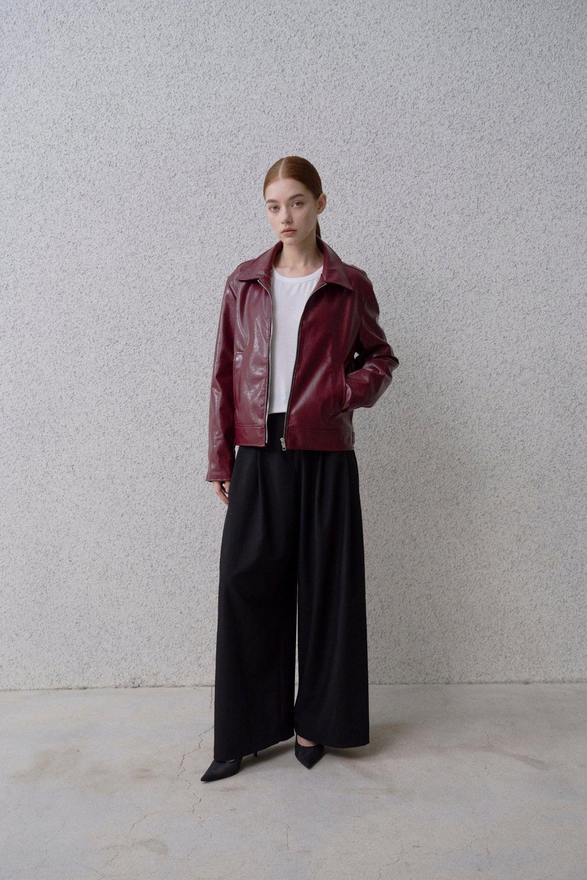 Woman wearing burgundy leather jacket, white top, wide-leg black pants, and black pointed heels against gray wall