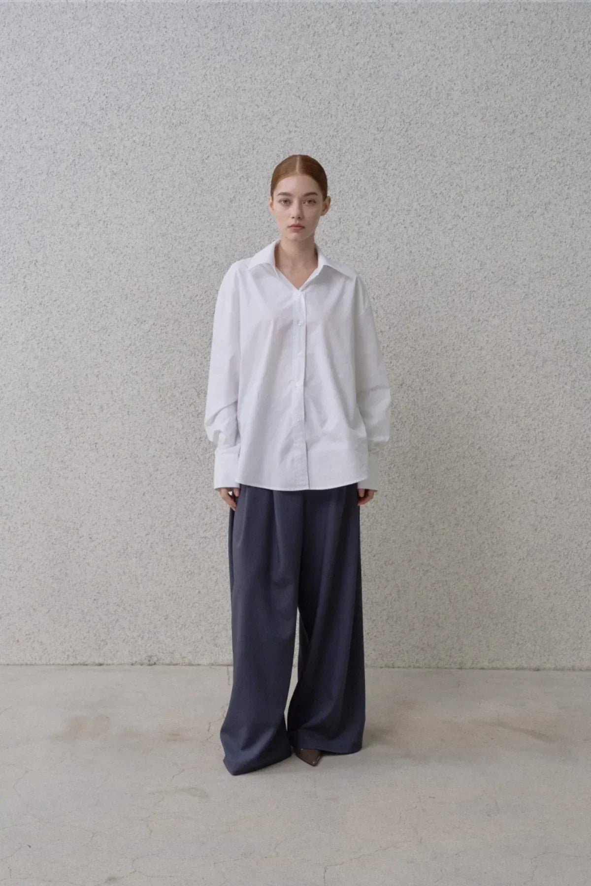 Model wearing a white oversized button-up shirt and wide-fit navy blue pants against a gray textured wall