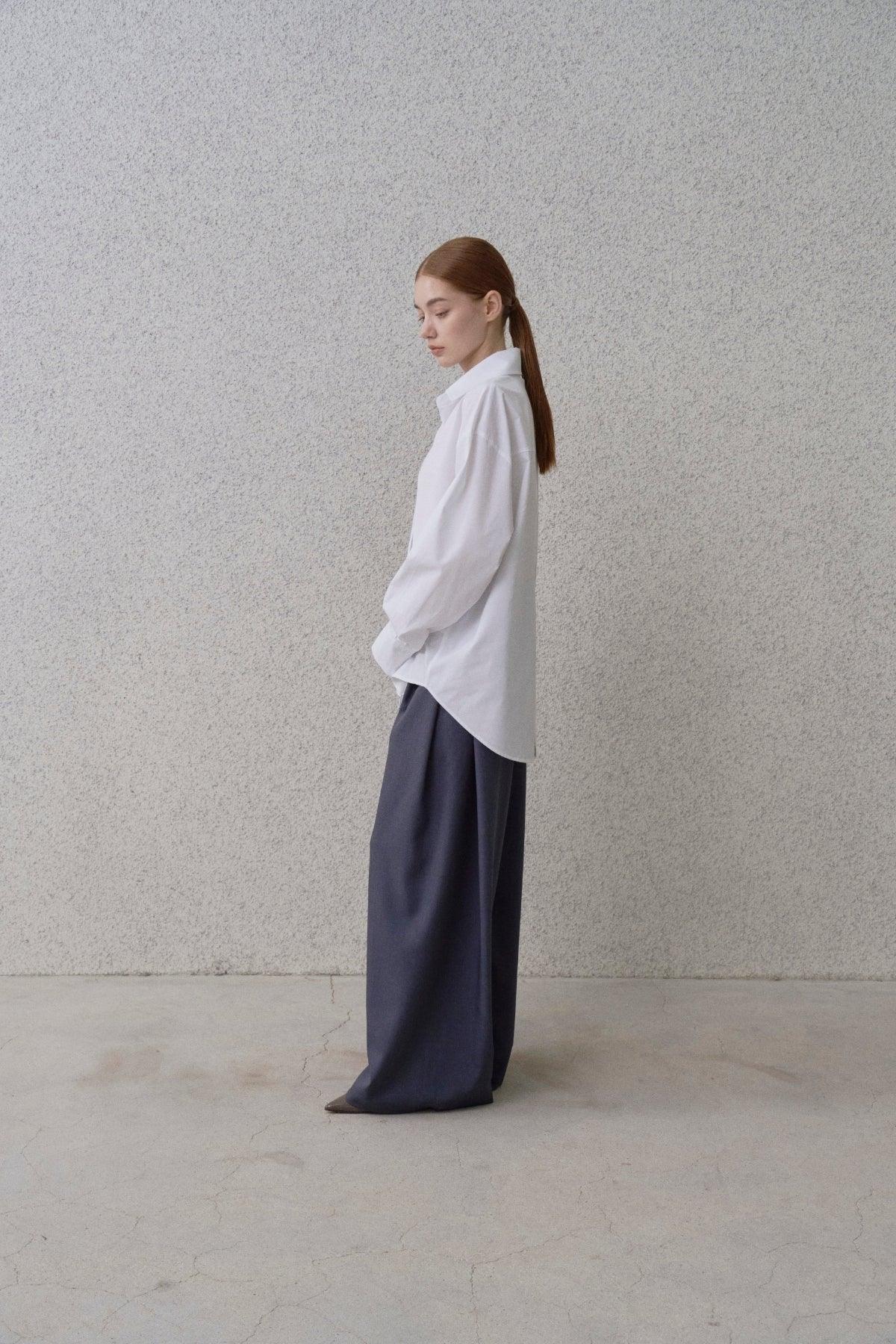 Side view of a woman wearing a loose white shirt and wide-fit navy blue pants against a gray textured wall