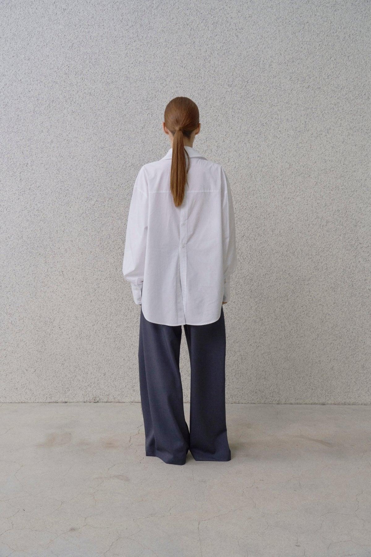 Rear view of woman with ponytail wearing white button-back shirt and wide-fit blue pants against gray wall