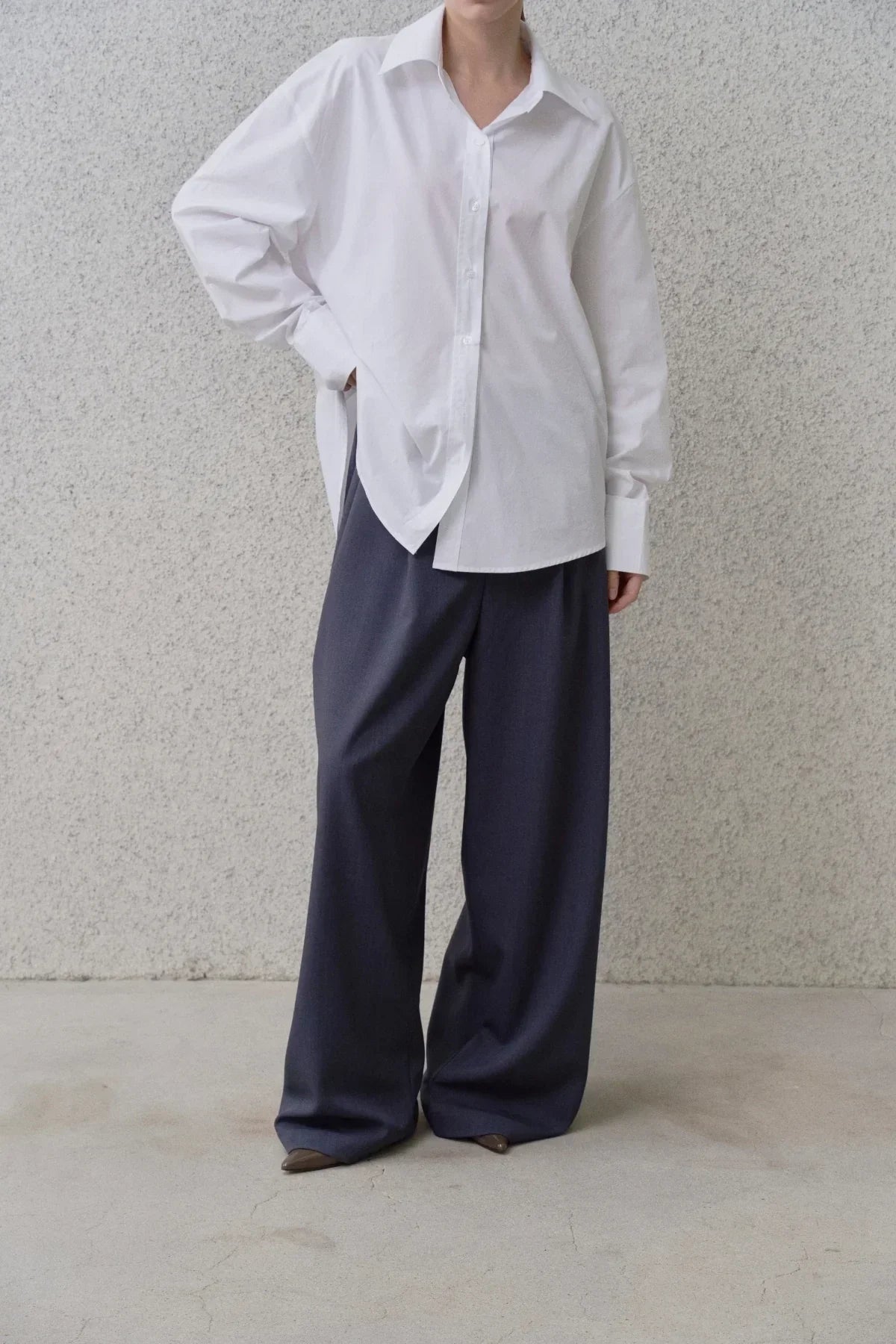 Model wearing wide-fit navy blue pants and a loose white button-up shirt against a textured gray wall
