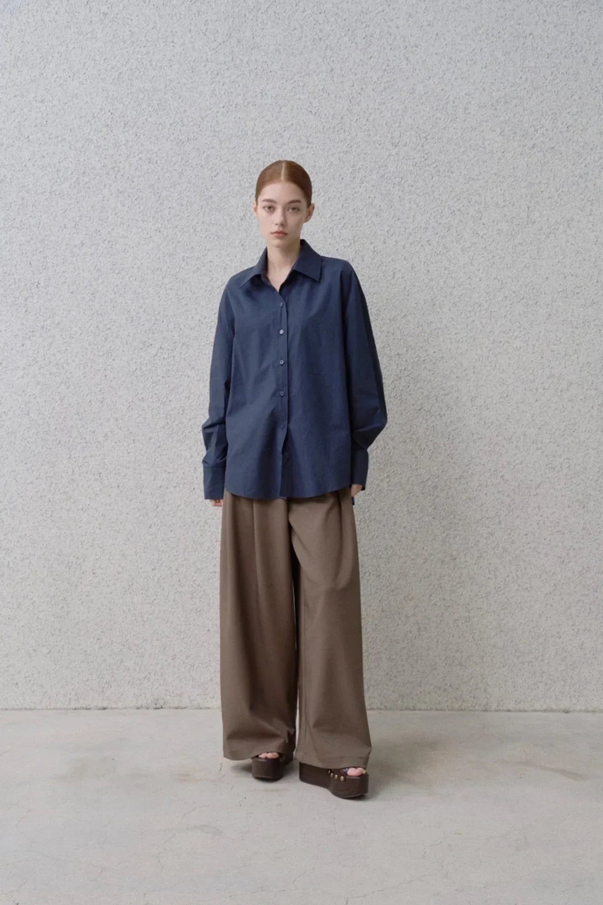 Model wearing navy button-up shirt and wide fit brown pants with platform sandals against gray wall