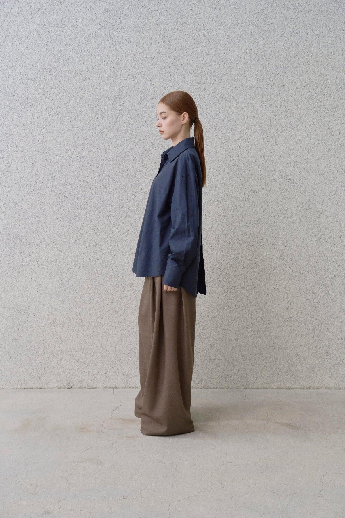 Side view of a woman in a navy blue oversized shirt and wide fit brown pants against a textured gray wall