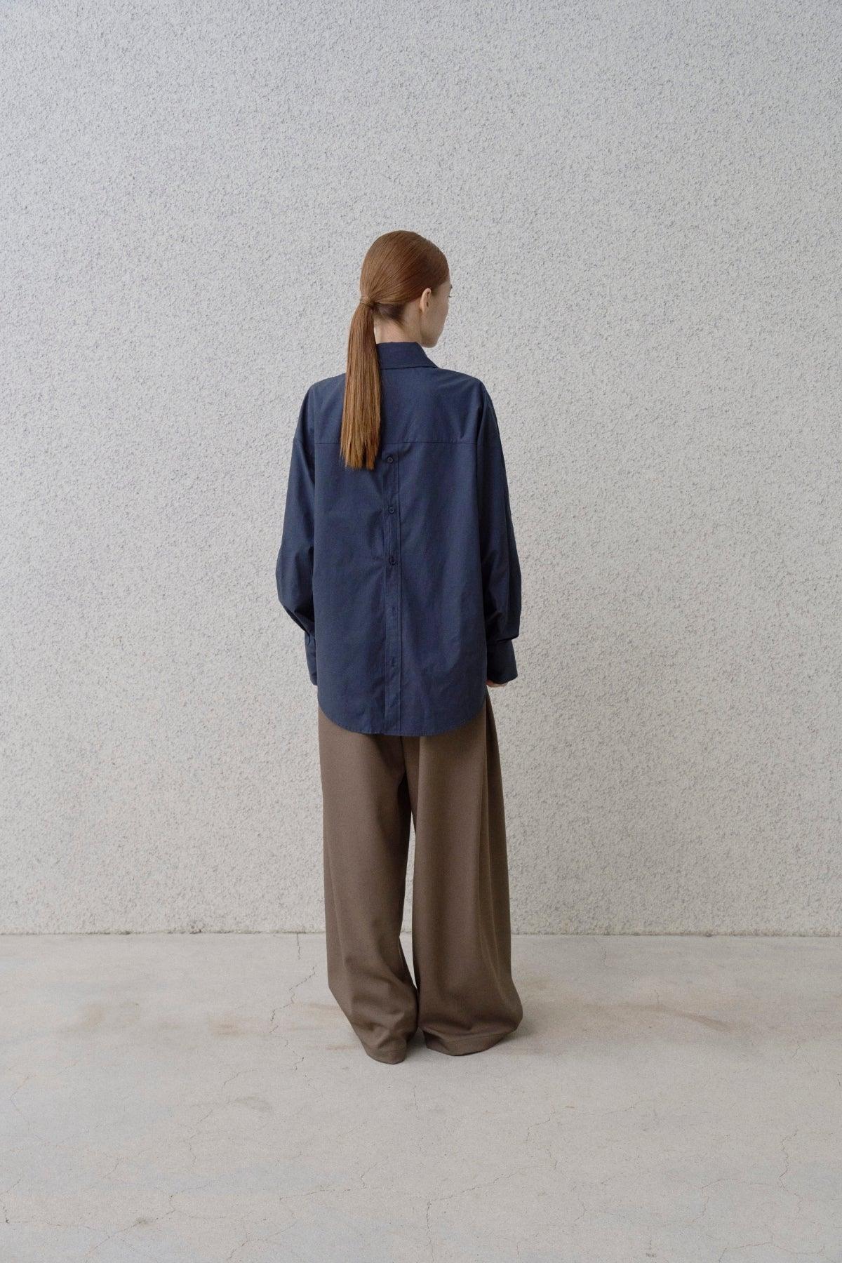 Woman with ponytail wearing oversized navy blue buttoned shirt and wide fit brown pants against textured gray wall