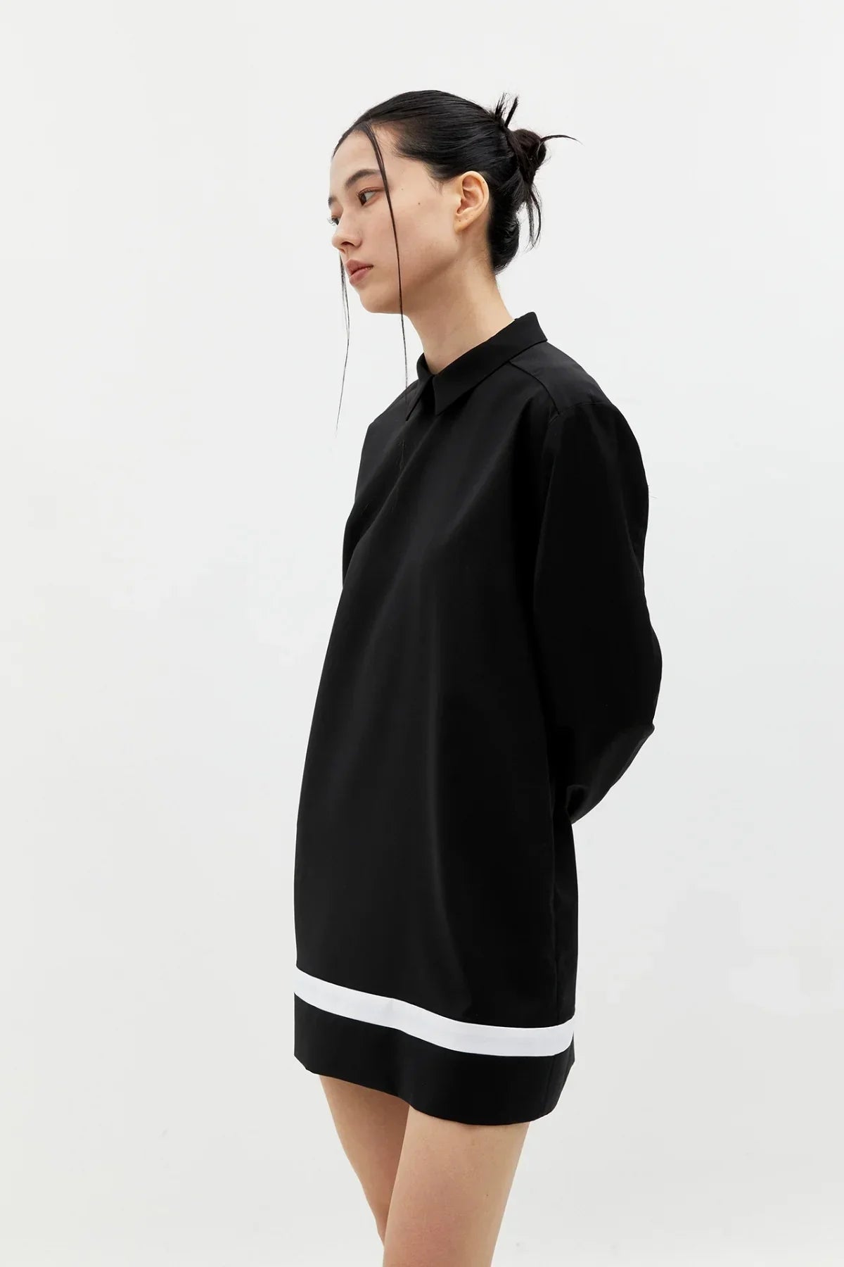 Side view of woman wearing black buttonless shirt dress with white stripe hem against white background