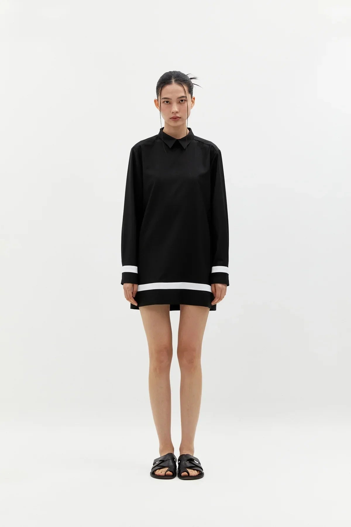 Model wearing buttonless black long-sleeve shirt dress with white trim and black slip-on sandals