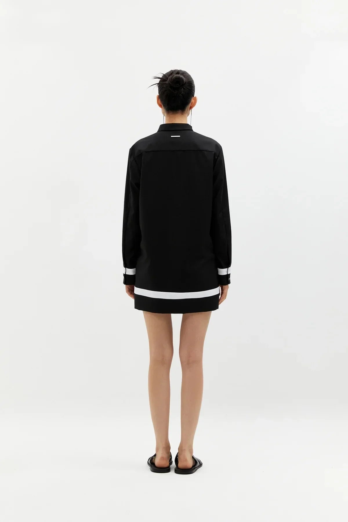 Woman wearing black buttonless shirt dress with white stripes on cuffs and hem, standing against a white background