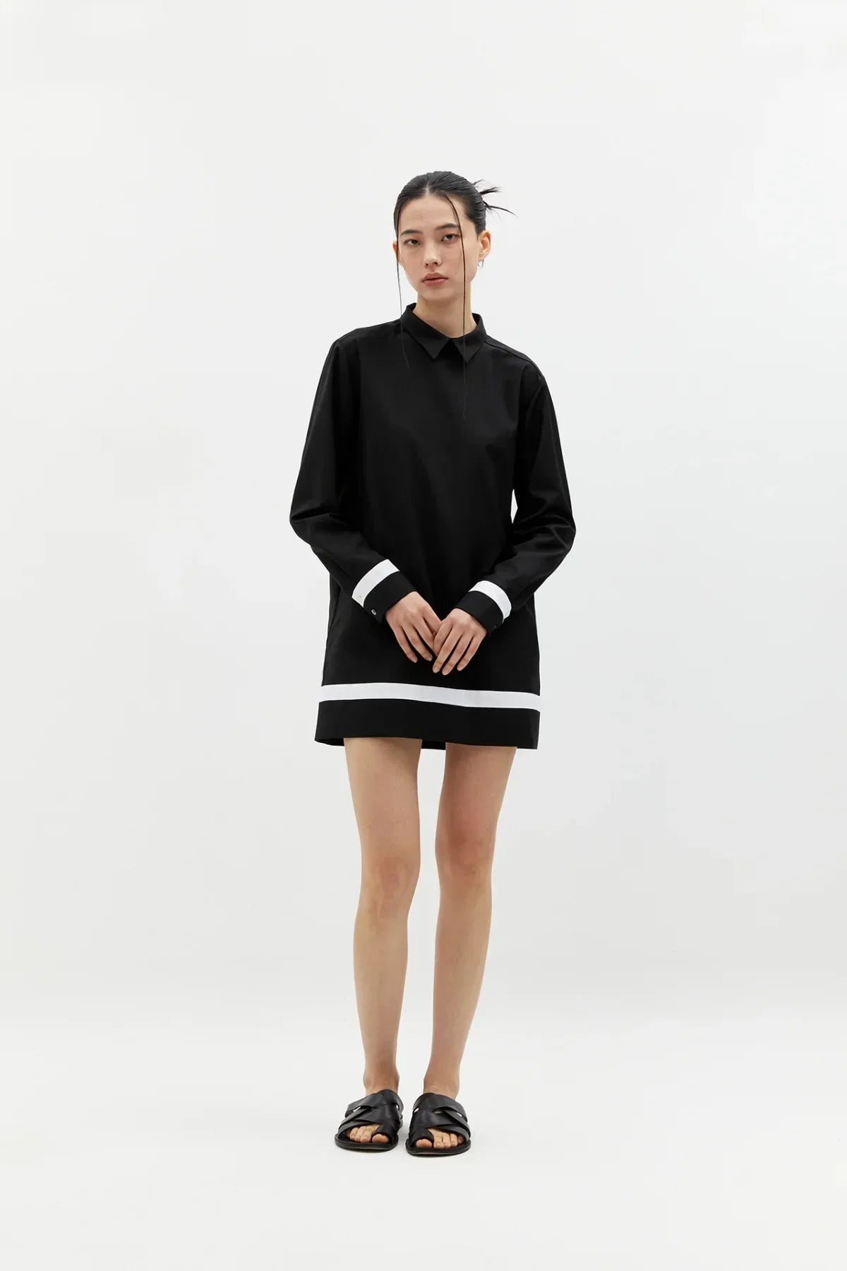 Model wearing a black buttonless long-sleeve shirt dress with white cuffs and hemline, styled with black sandals on white background